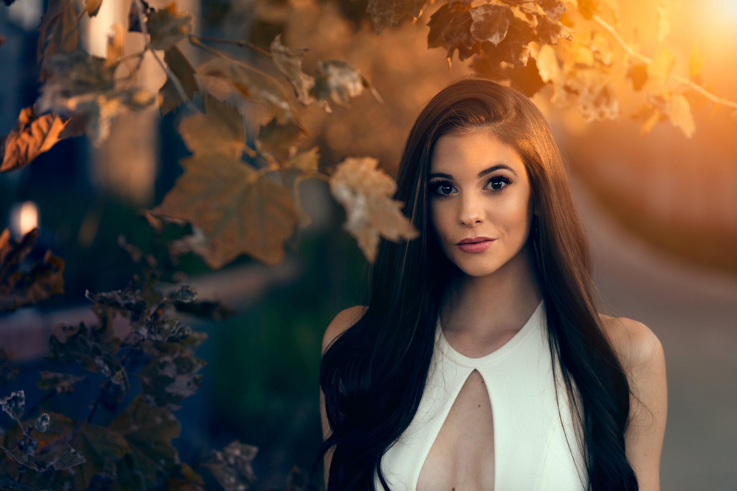 A young woman with long dark hair wearing a sleeveless white top stands outdoors among autumn leaves with warm sunlight in the background.