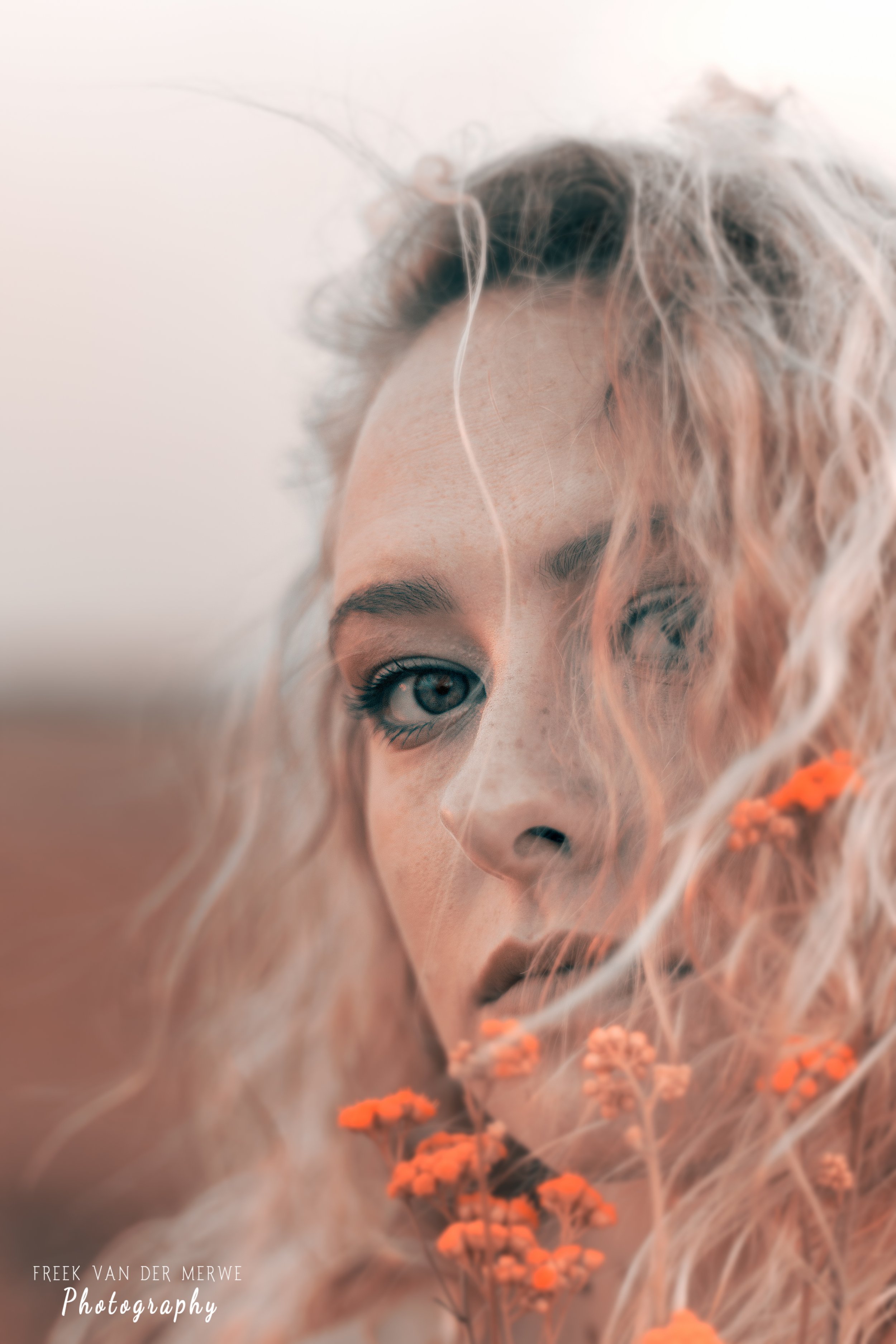 Close-up of a woman with curly blonde hair, one eye closed, holding orange flowers near her face.