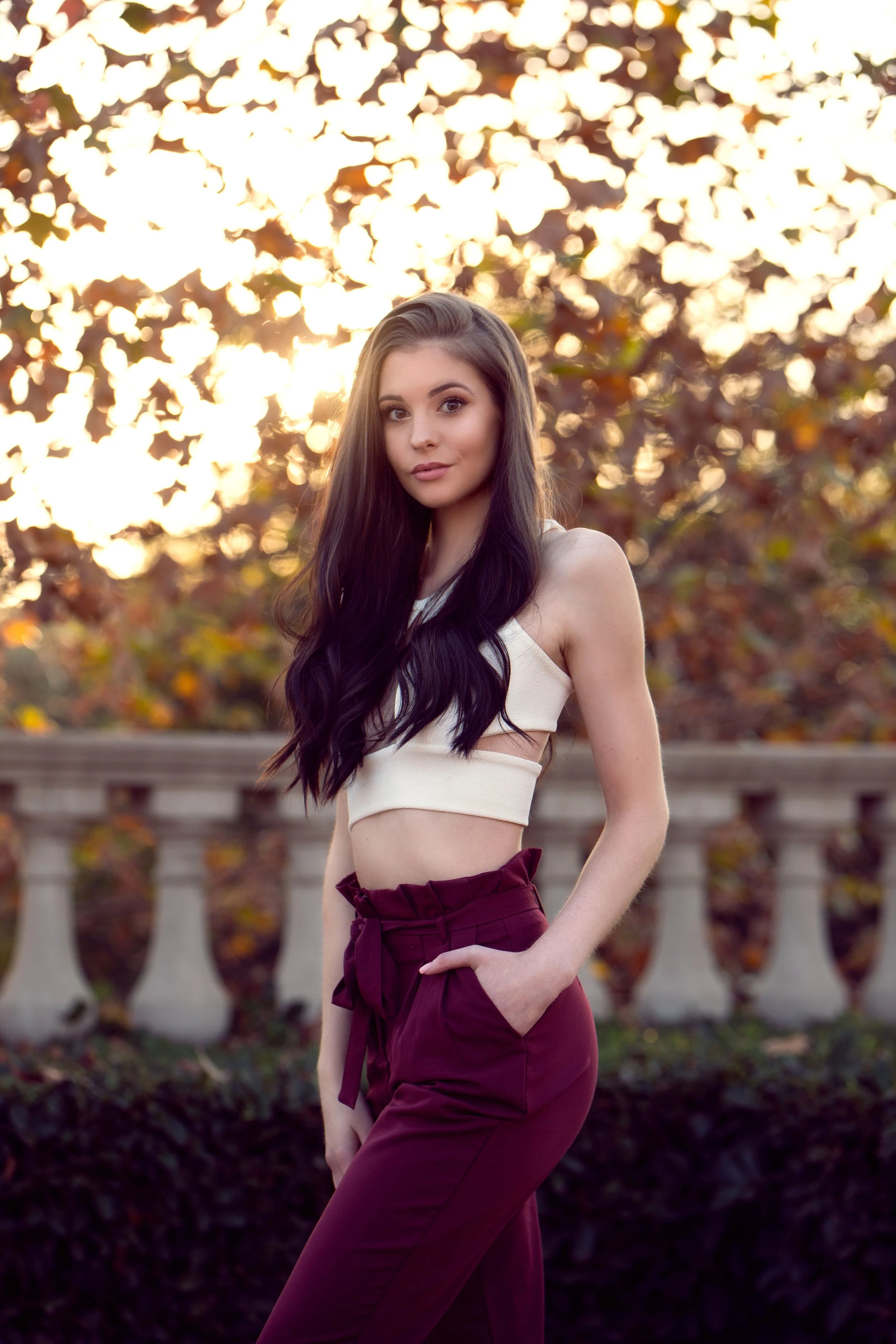 A young woman with long dark hair wearing a sleeveless beige crop top and high-waisted maroon pants, standing outdoors during sunset with trees and a stone balustrade in the background.