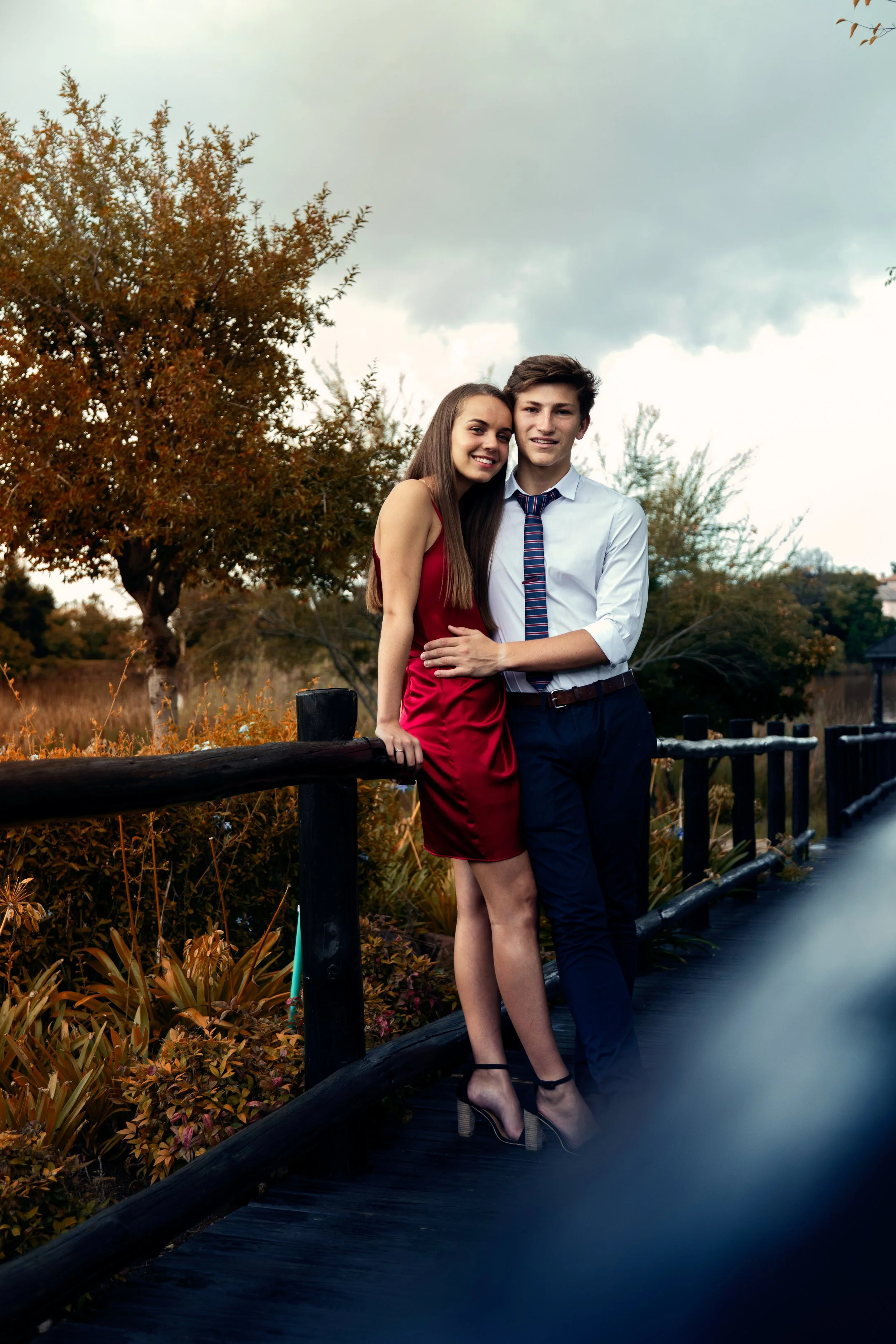 A young couple stands together on a wooden bridge outdoors, smiling. The woman wears a red dress and black high-heeled shoes, and the man wears a white shirt with rolled-up sleeves, a tie, and dark pants. Behind them are trees with autumn-colored lea