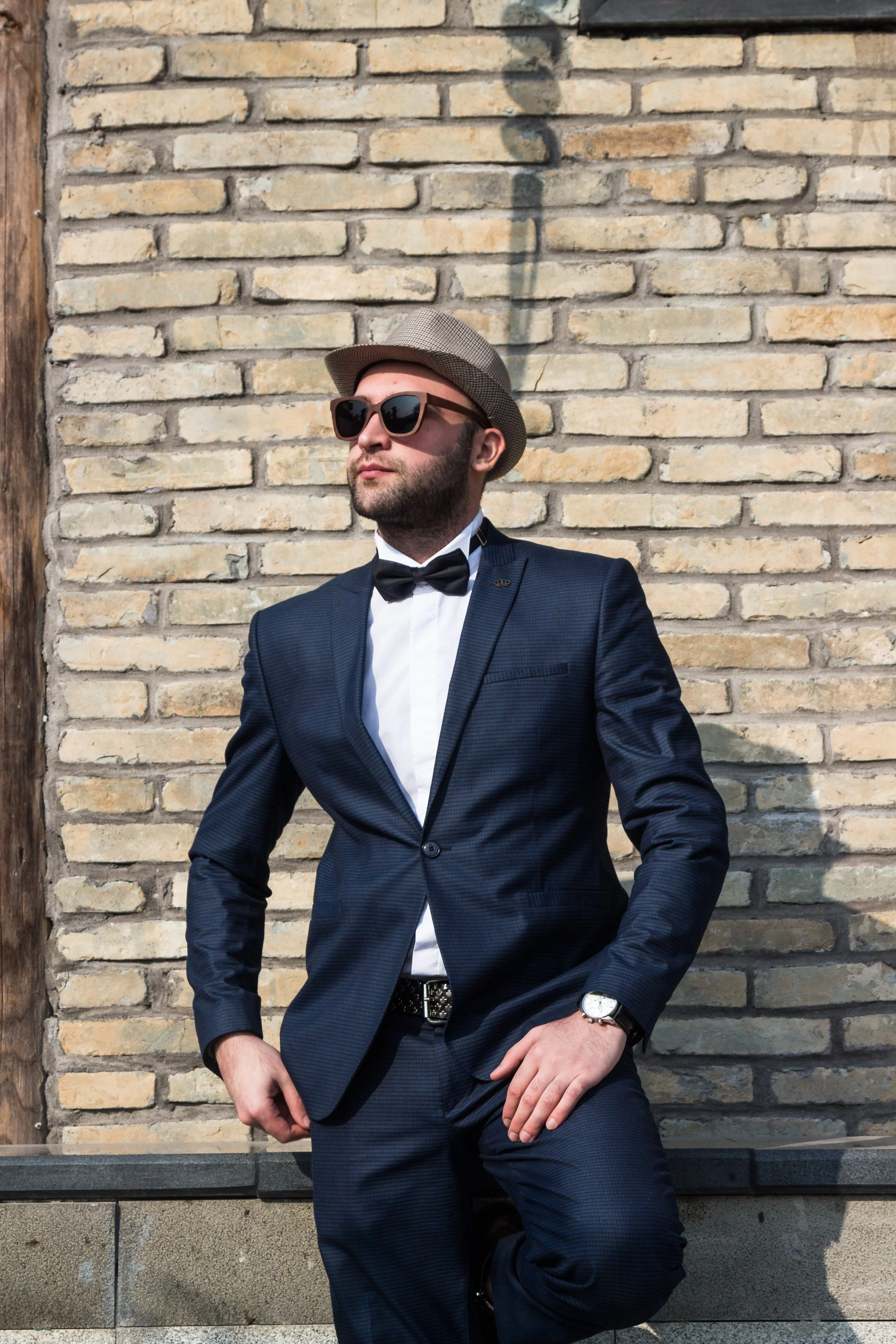 A man in a navy tuxedo with a bow tie, wearing sunglasses, a watch, and a hat, posing in front of a brick wall.