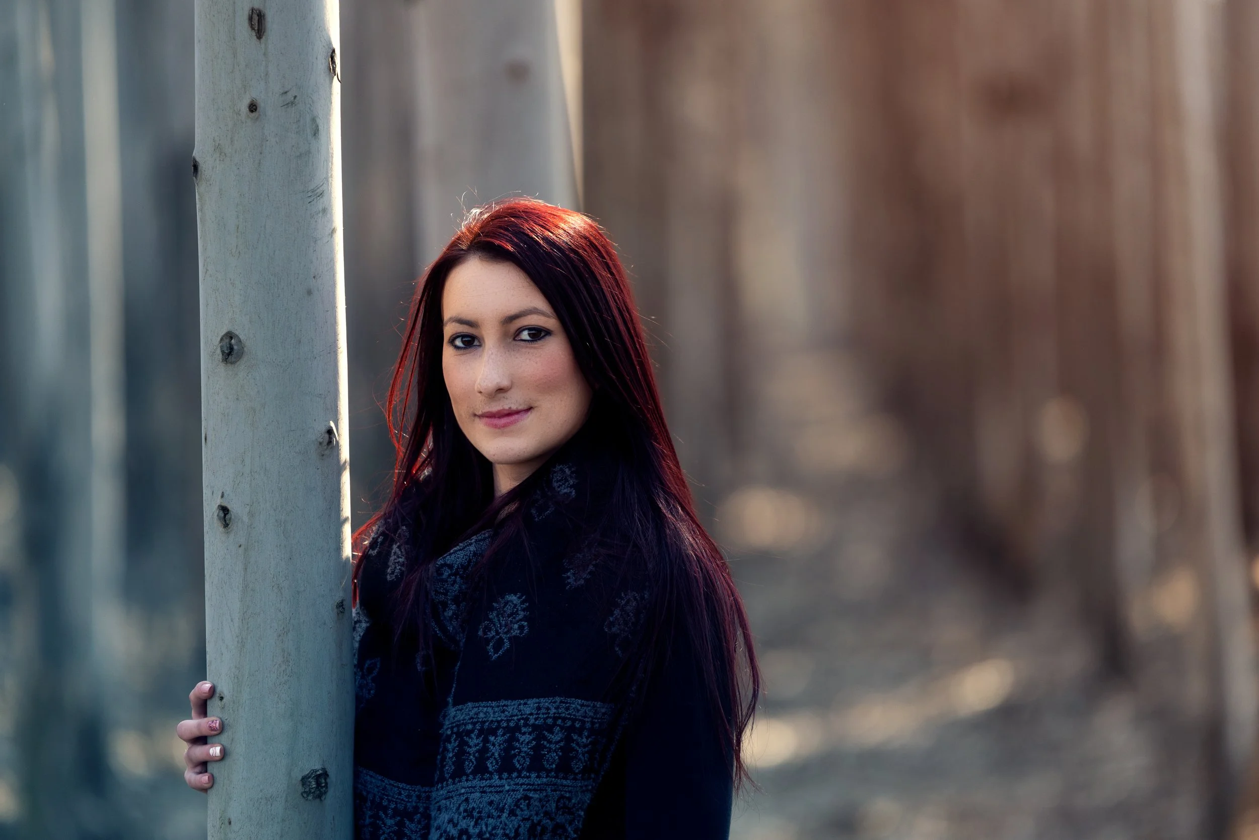 Young woman with long dark red hair holding onto a tree trunk outdoors in a forest setting during sunset.
