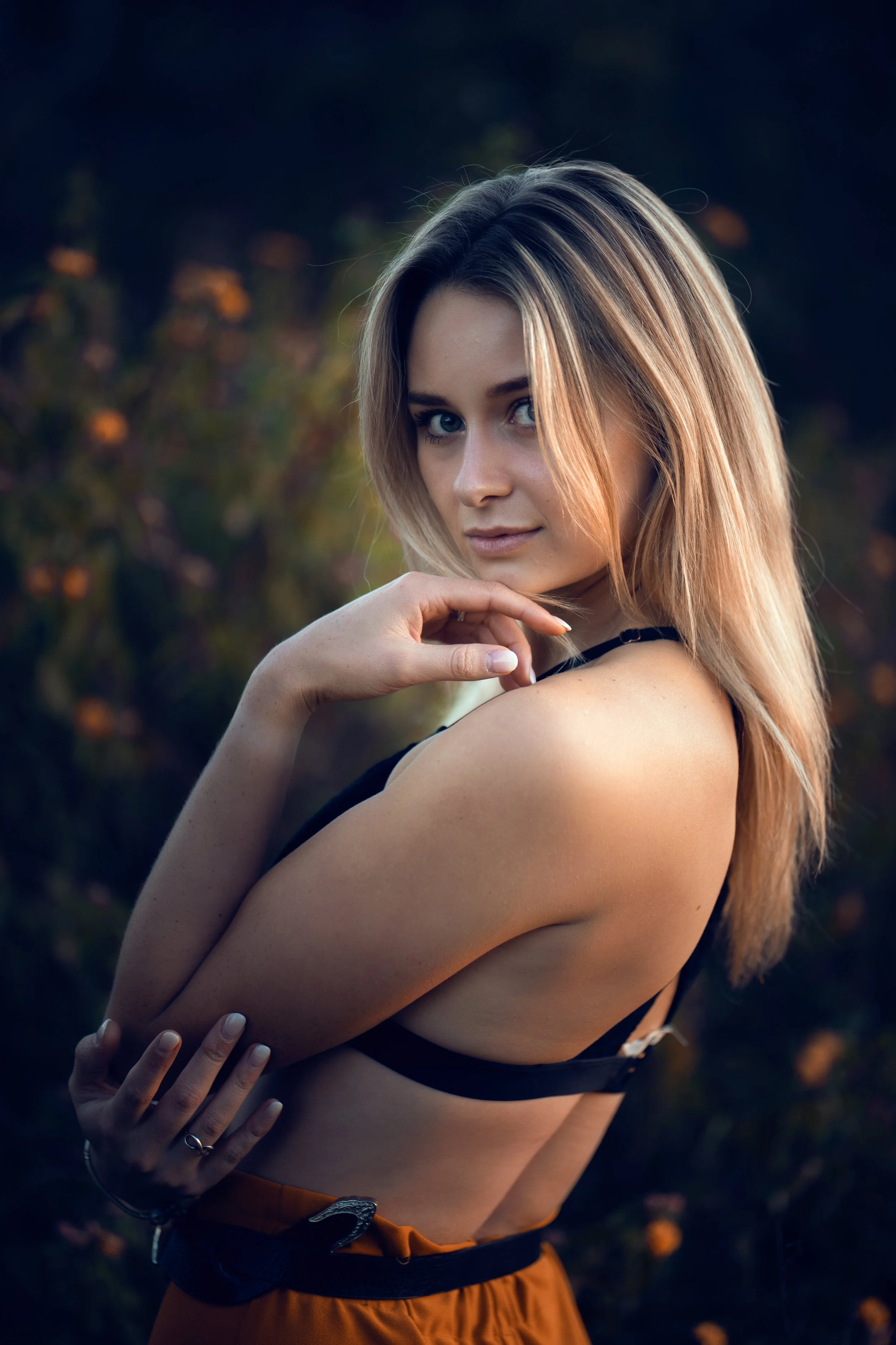 A young woman with blonde hair and blue eyes standing outdoors during late afternoon or early evening, wearing a black top and orange pants, with a blurred natural background.