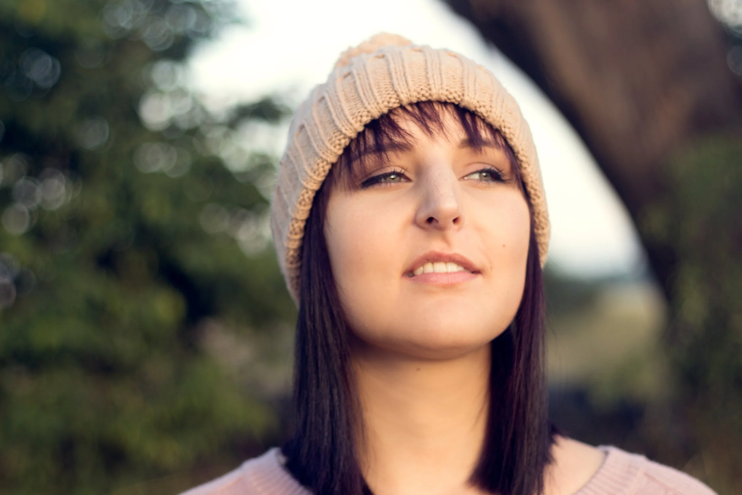 A young woman outdoors wearing a beige knit beanie, with dark hair, light makeup, and a slight smile.