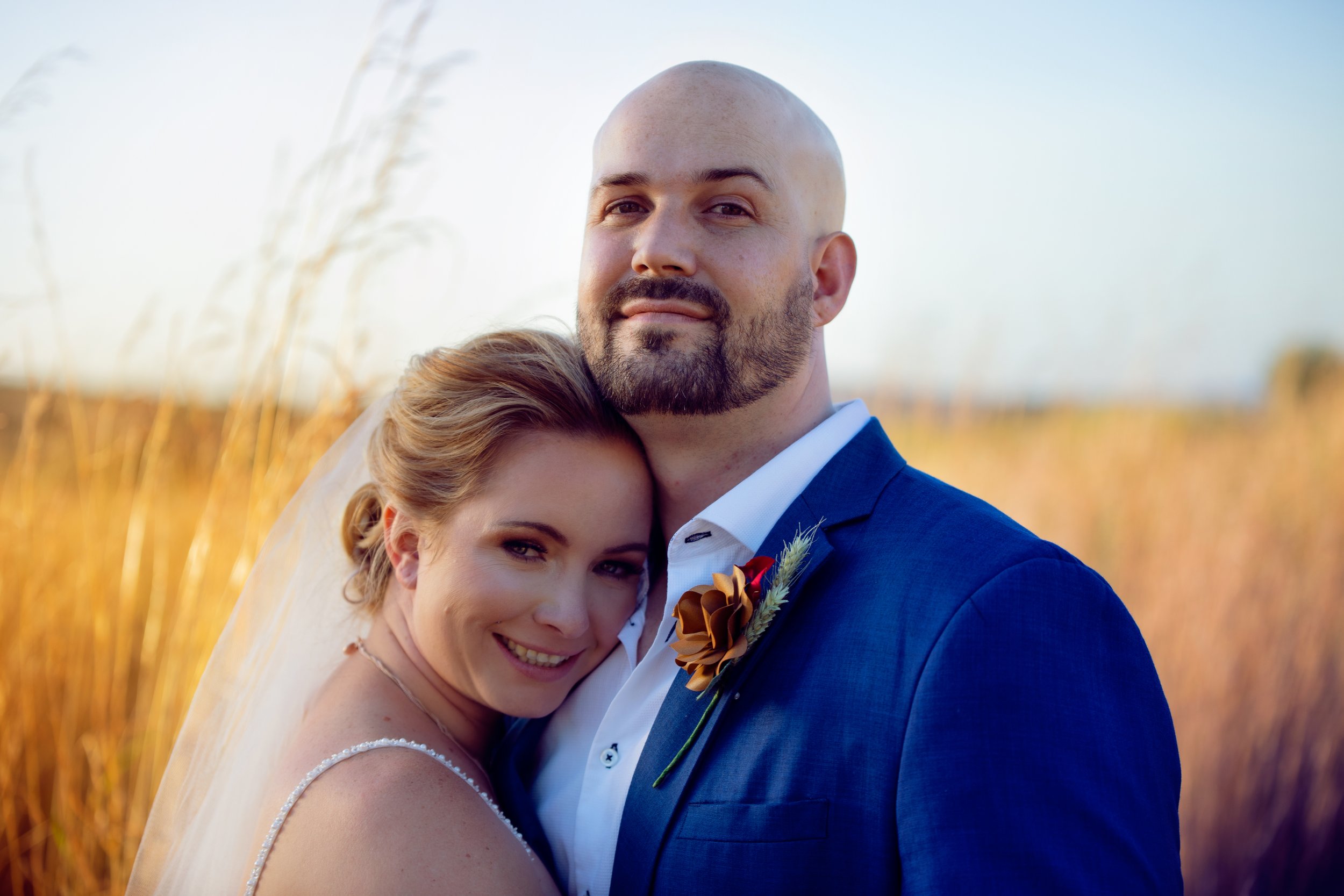 A newlywed couple embraces outdoors in a field of tall golden grass during sunset. The woman smiles, resting her head on the man's shoulder, who is dressed in a blue suit with a boutonnière.