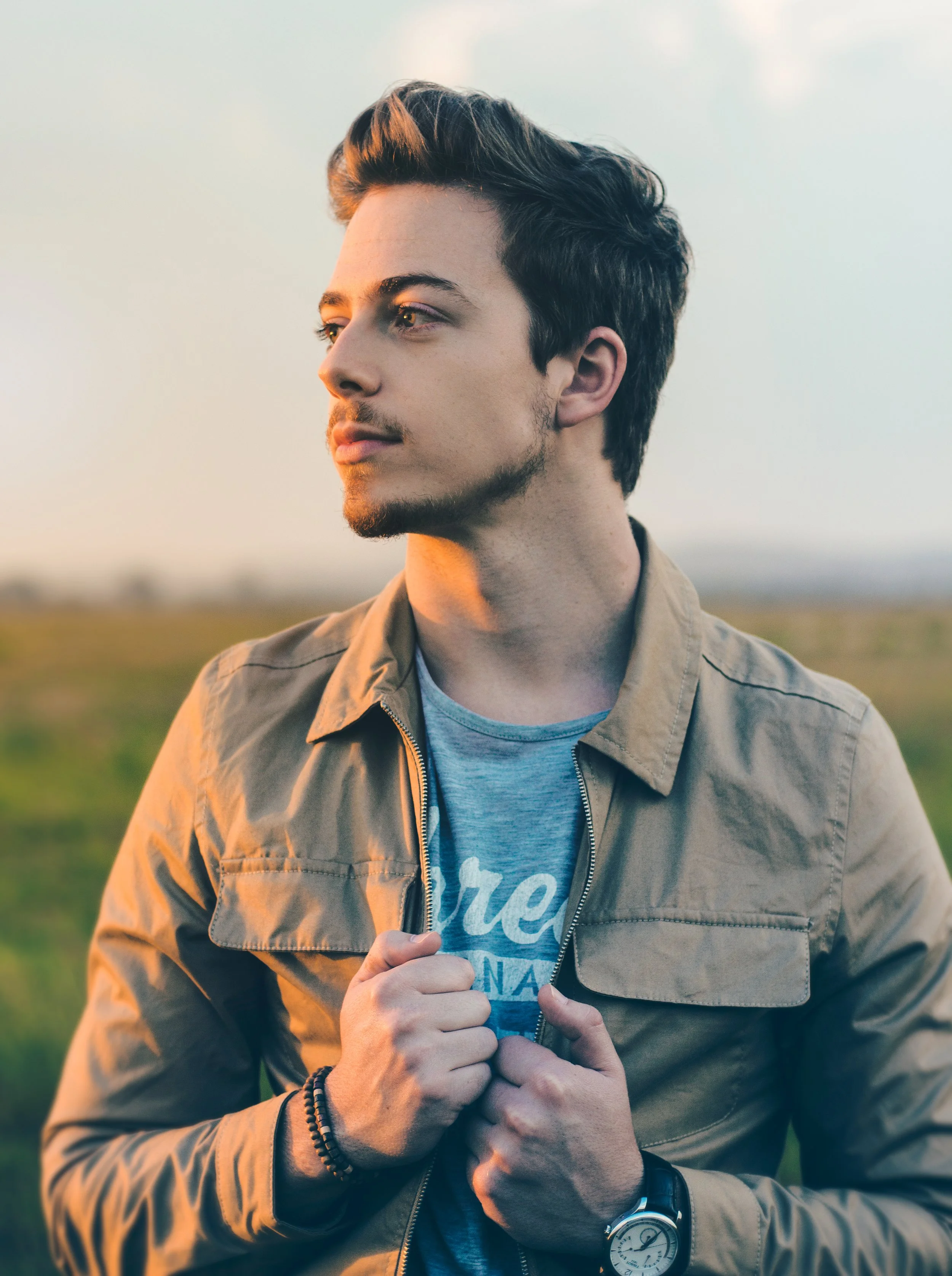 A young man with styled hair and facial hair, wearing a tan jacket over a gray T-shirt, standing outdoors with a blurred landscape in the background during sunset.