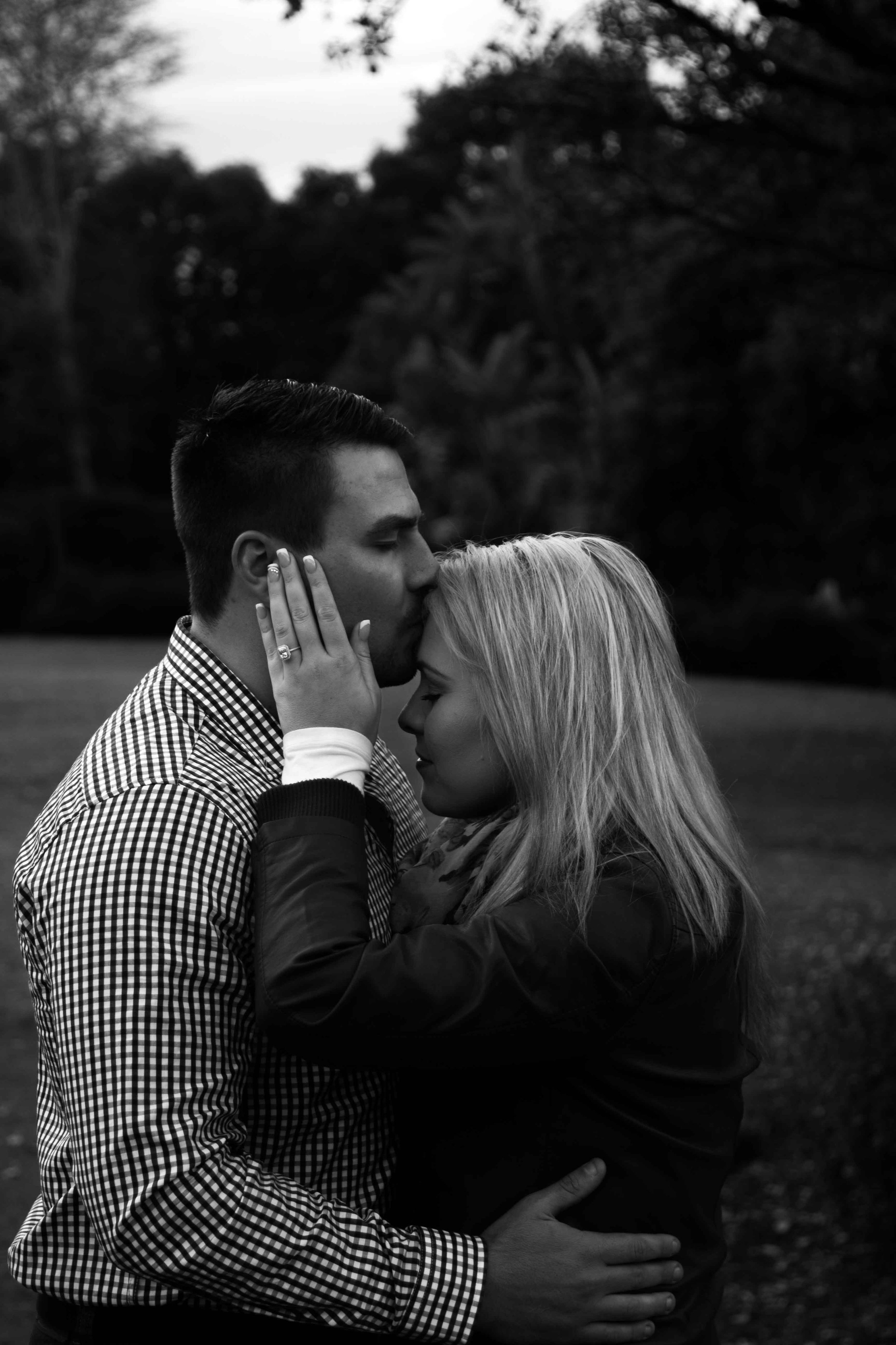 A black and white photo of a loving couple embracing outdoors, with the man kissing the woman's forehead and their eyes closed.