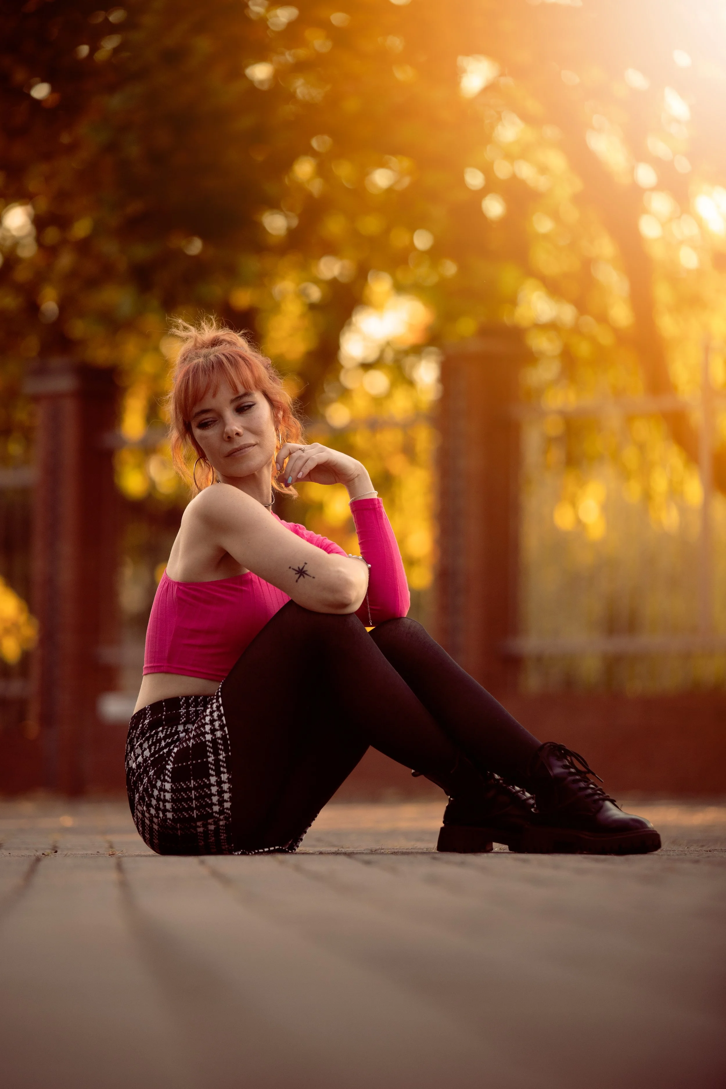A young woman with red hair and a star tattoo on her arm sitting on a wooden deck outdoors during sunset, wearing a pink sleeveless top, black leggings, and black lace-up boots, with trees and a brick fence in the background.