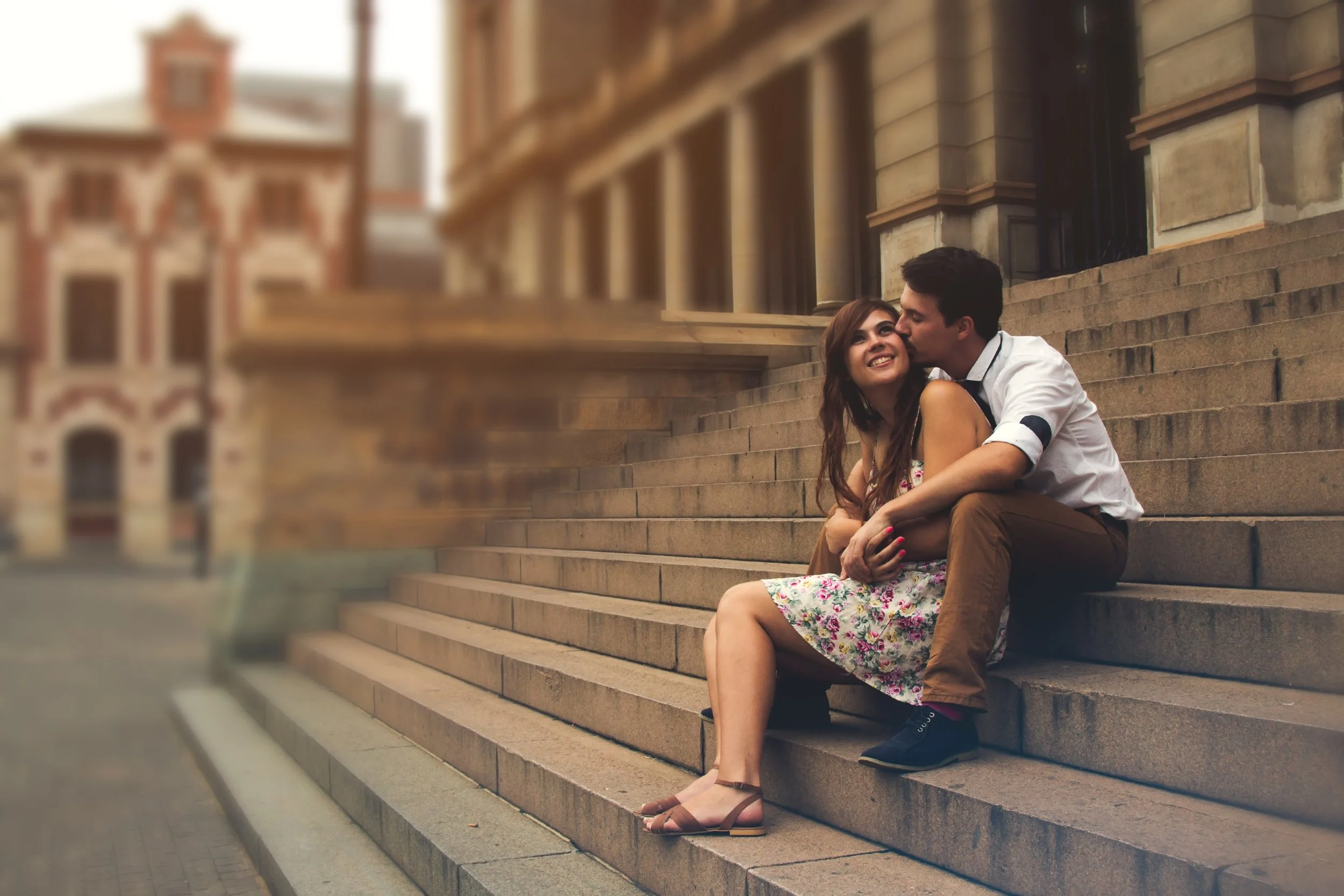 A young couple sitting on stone steps outside a historic building, with the man kissing the woman's cheek and both smiling, during daytime.