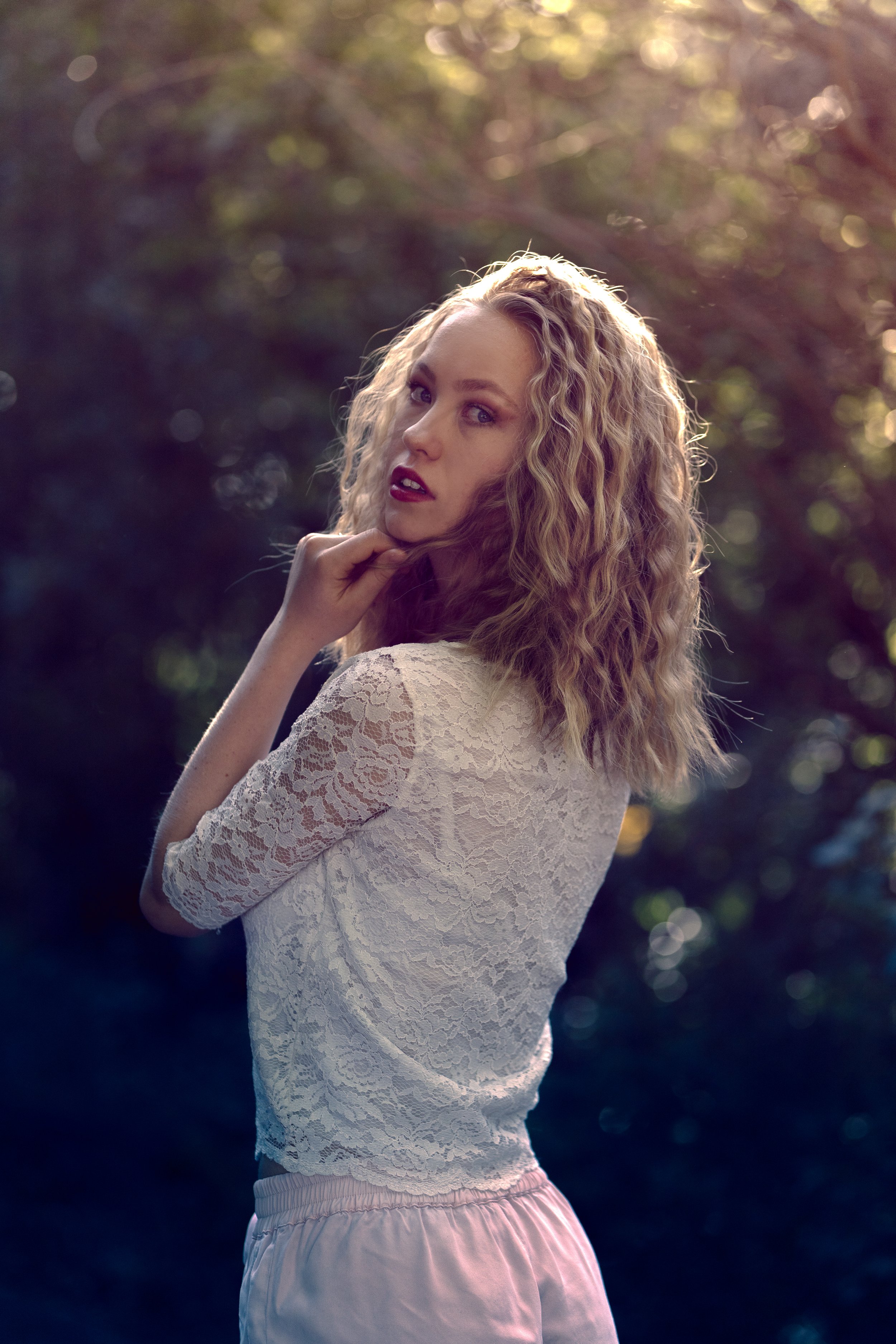 A woman with curly blonde hair standing outdoors, wearing a white lace top and pink shorts, with her hand gently touching her chin, looking at the camera.