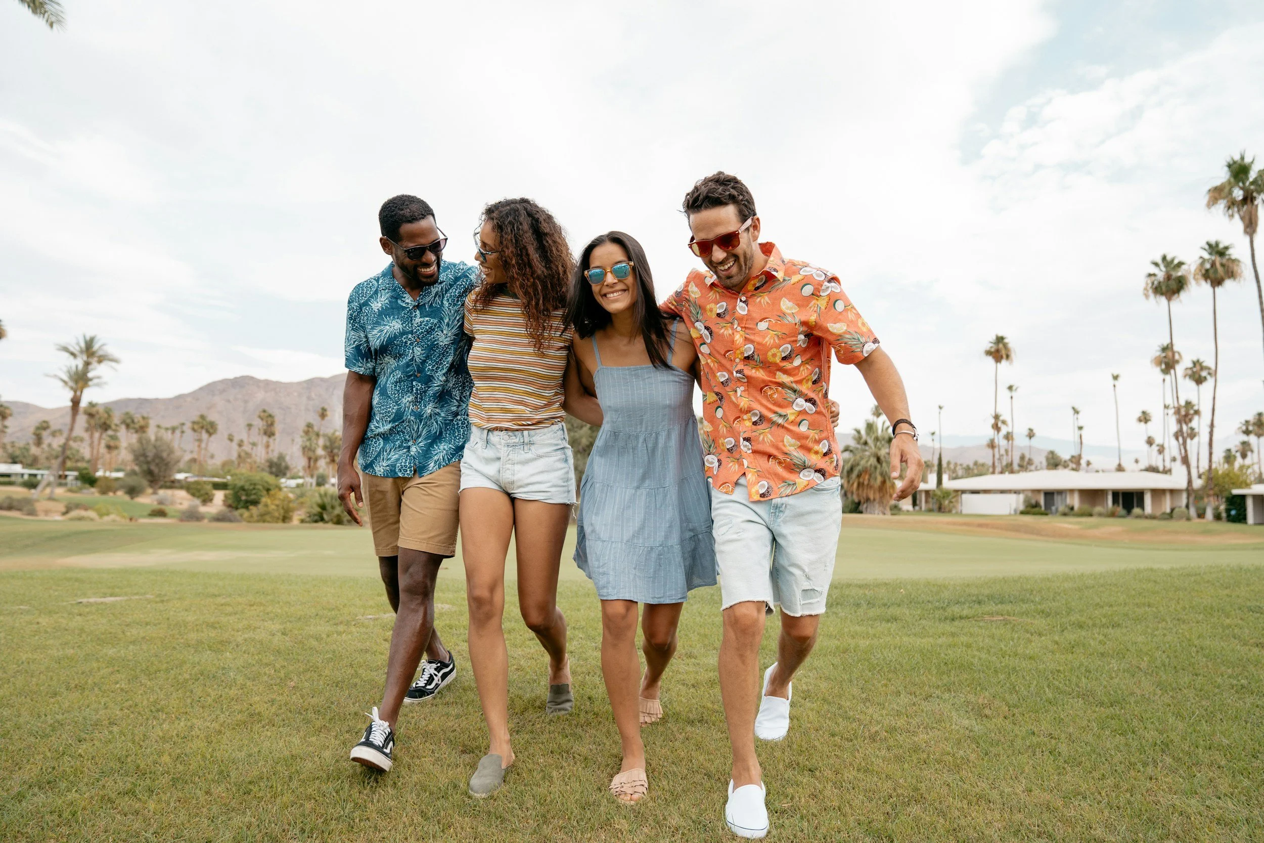 A group of four friends walking together on a grassy field in Orlando, Florida in a sunny outdoor setting with palm trees in the background, smiling and enjoying each other's company.