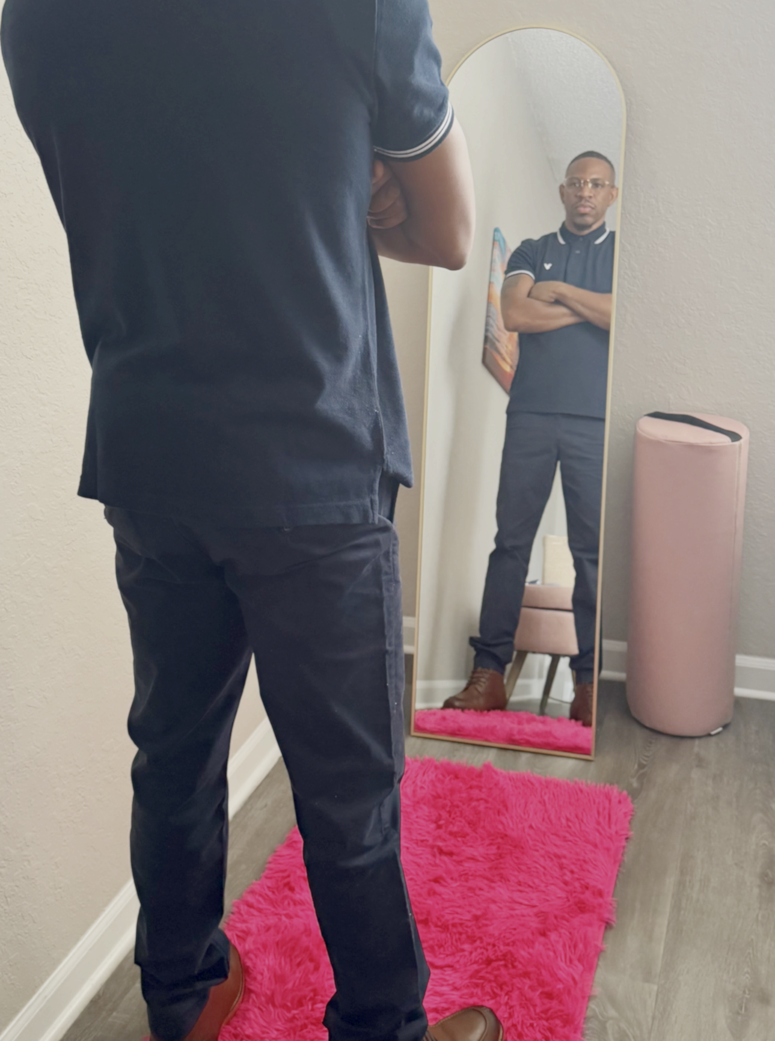 Coach Howard standing in front of a full-length mirror with arms crossed, wearing navy blue pants and polo shirt, and brown shoes, looking at the reflection in a massage therapy room with a pink fluffy rug, and a pink bolster pillow cushion.