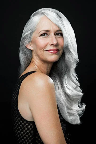 A portrait of a mature milf cougar woman with sleek long, shimmering white grey silver hair, wearing a black sleeveless top, and smiling slightly against a black background.