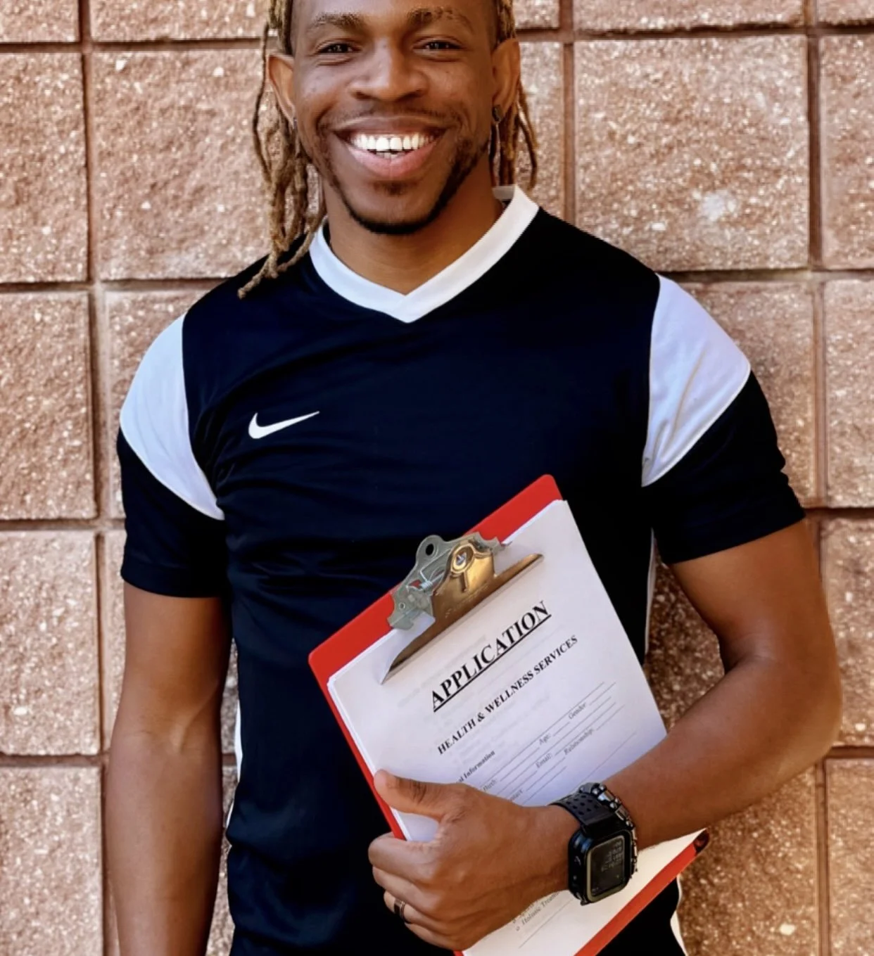 A strong handsome smiling black man holding an application form for health and wellness services in front of a brick wall.