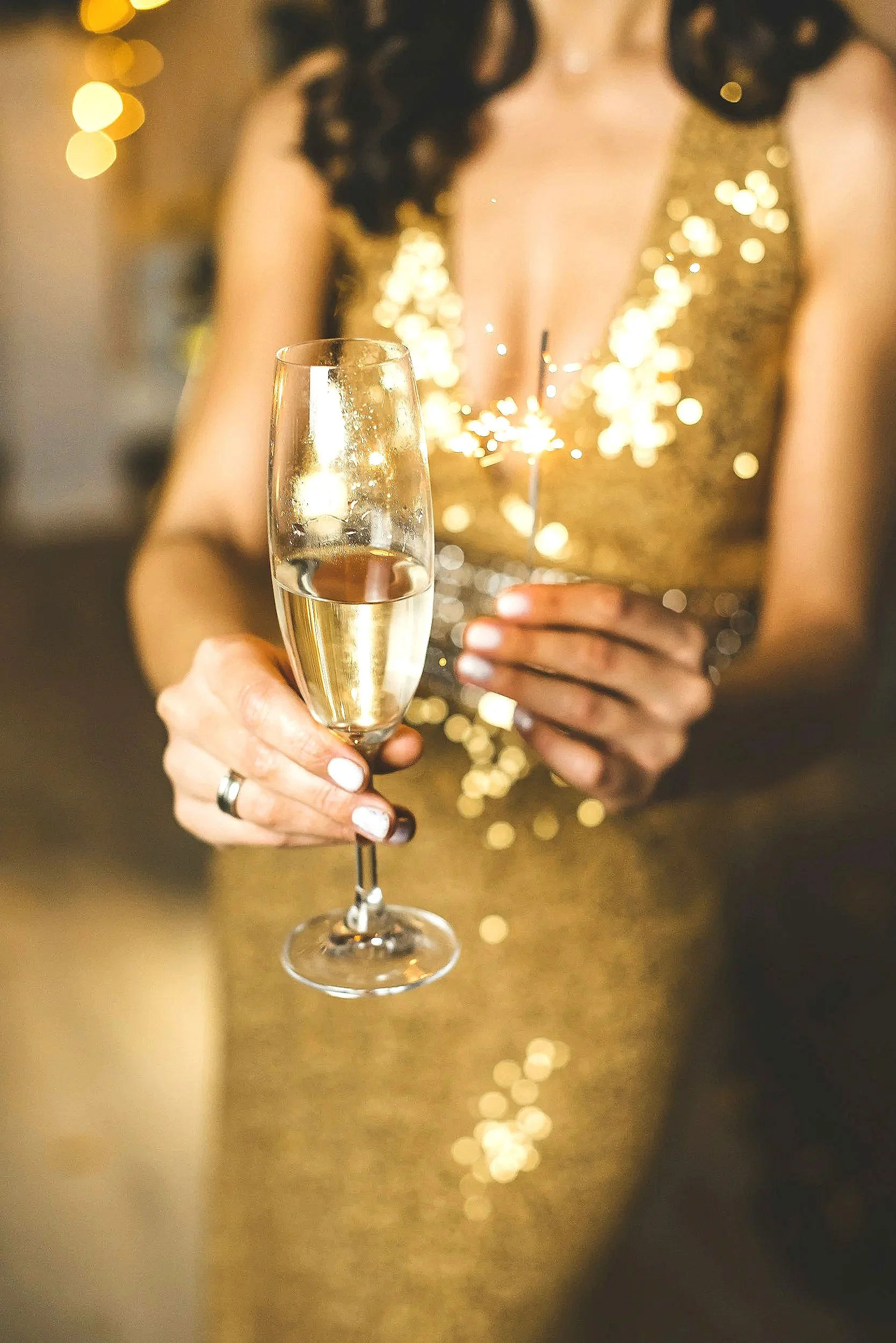 A beautiful elegant gorgeous blonde brunette woman in a gold sequined dress holds a sparkler and a glass of champagne during a health and wellness celebration.