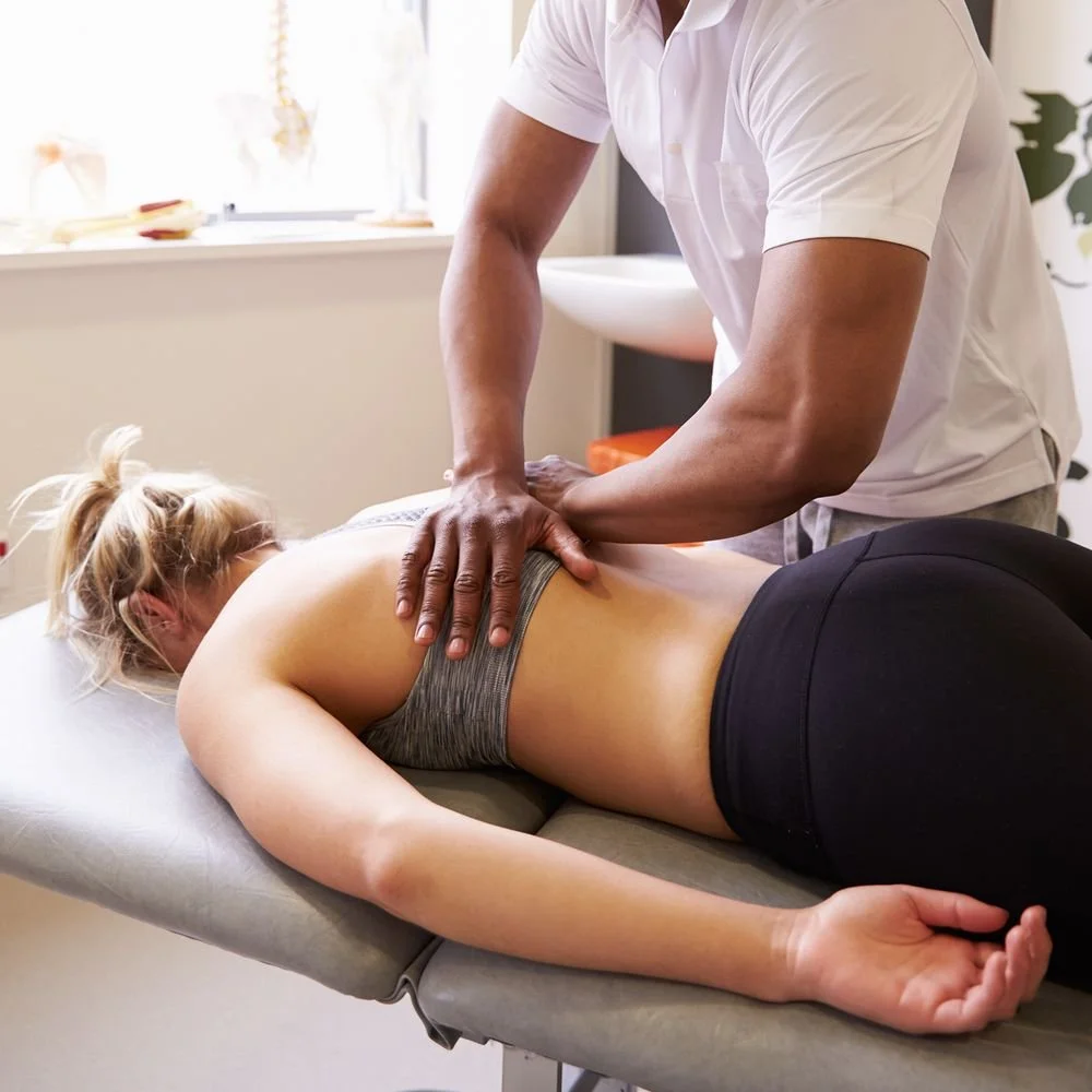 Big black man handsome strong masculine male massage therapist performing deep tissue techniques on a resting sleeping beautiful woman lying face down on a treatment table in a bright clinical setting.
