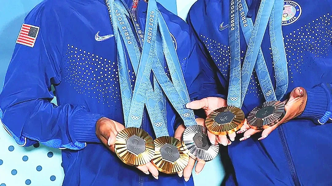 Two athletes wearing blue jackets with medals around their necks, holding gold and silver medals in their hands.