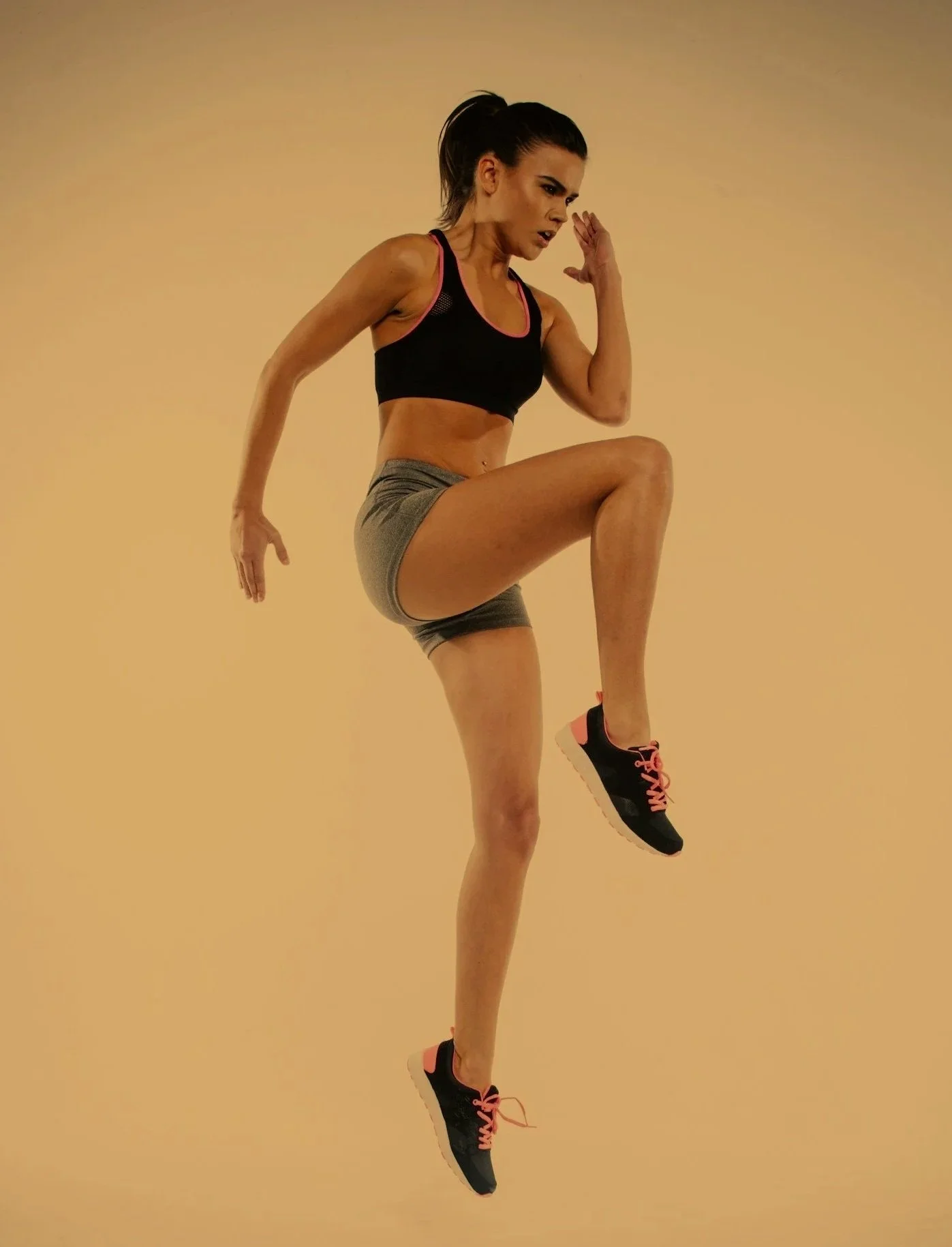 A young brunette or blonde teen woman in booty shorts, sports bra, sneakers and workout attire performing a high knee exercise against a plain beige background.