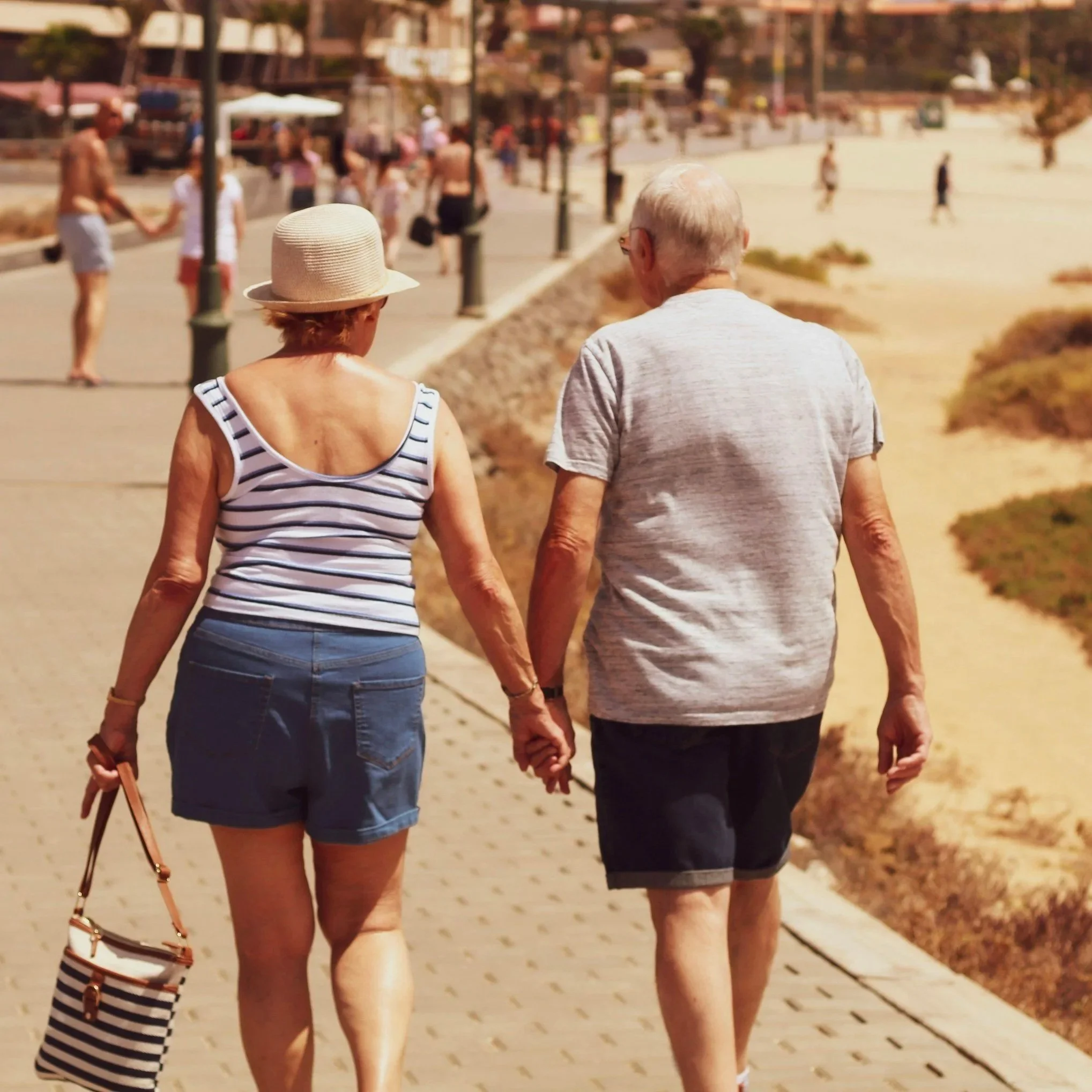 An elderly couple walking hand-in-hand along a paved pathway on a sunny day, with people and buildings in the background.