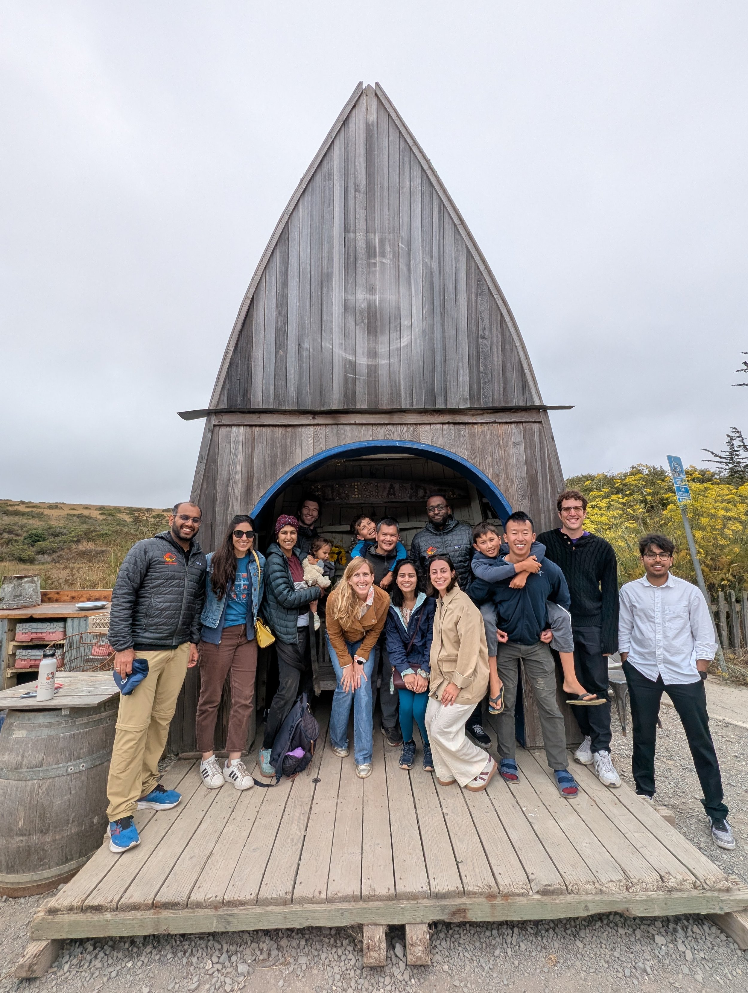 Group of people posing in front of a wooden building with a pointed roof and a large circular opening.