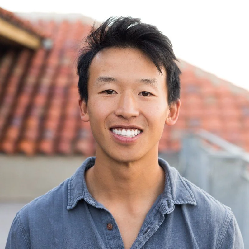 Young man smiling outdoors, wearing a blue button-up shirt, with a roof and cloudy sky in the background.