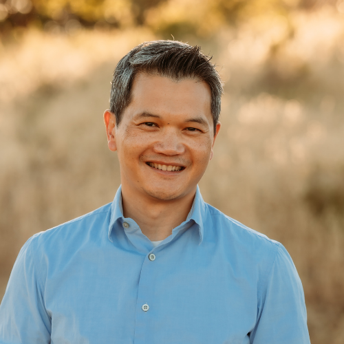 A smiling man in a light blue dress shirt standing outdoors with a blurred natural background.