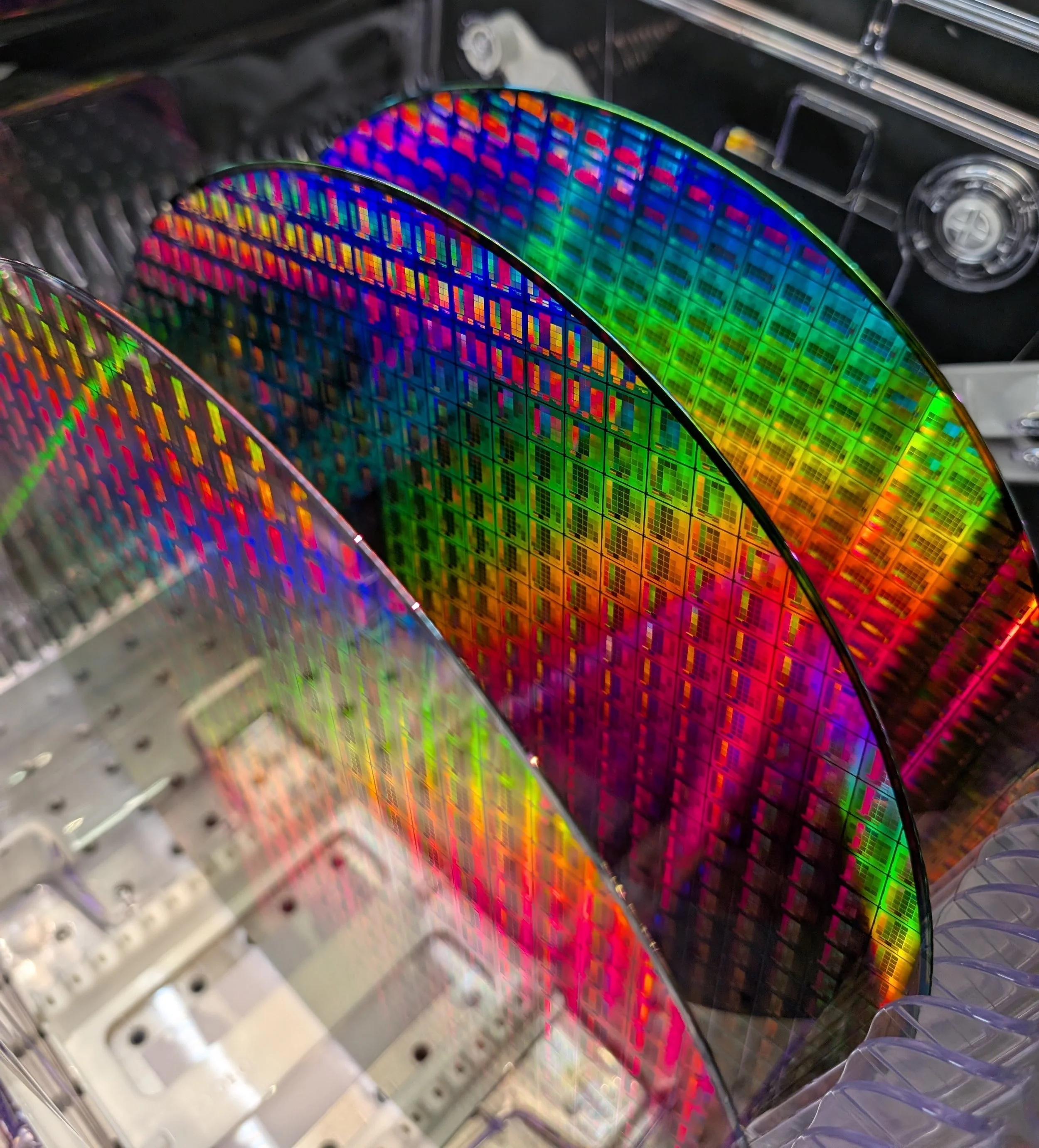 Close-up of multiple colorful nanophotonic wafers, showing bright, reflective surface with intricate circuitry and gold contacts, inside a plastic protective packaging.