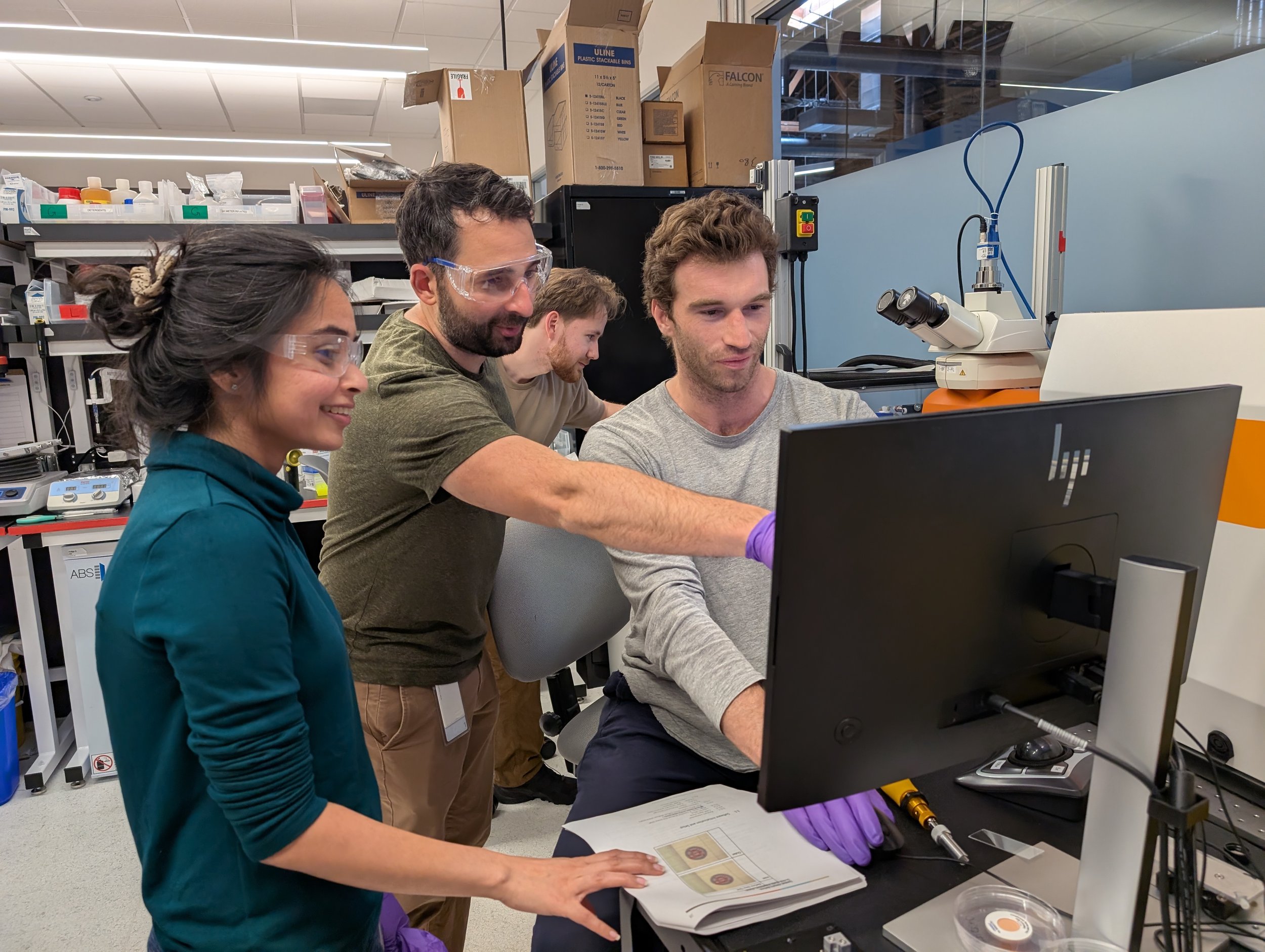 A group of scientists working in a laboratory with a microscope, computer, and various lab supplies, wearing safety glasses.