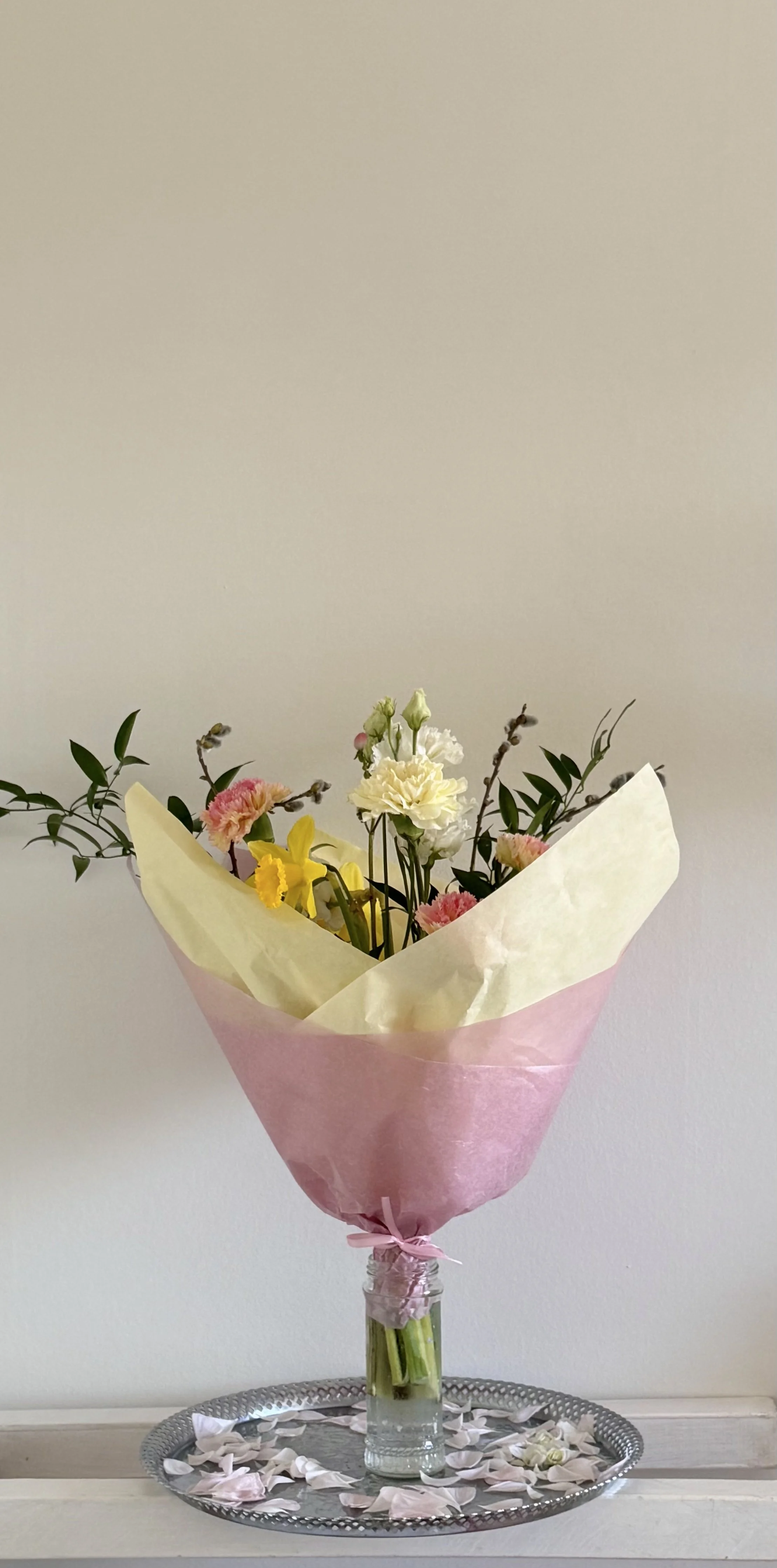 A bouquet of mixed flowers, including pink, white, and yellow blossoms, wrapped in pink and cream paper, in a glass jar, placed on a tray with scattered flower petals against a plain background.