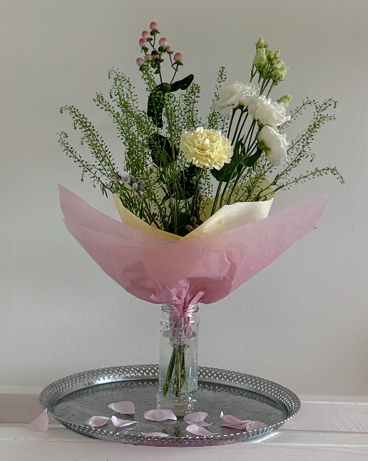 A bouquet of white, pink, and green flowers in a pink paper wrapping, placed in a glass vase on a silver tray with pink petals scattered on it.