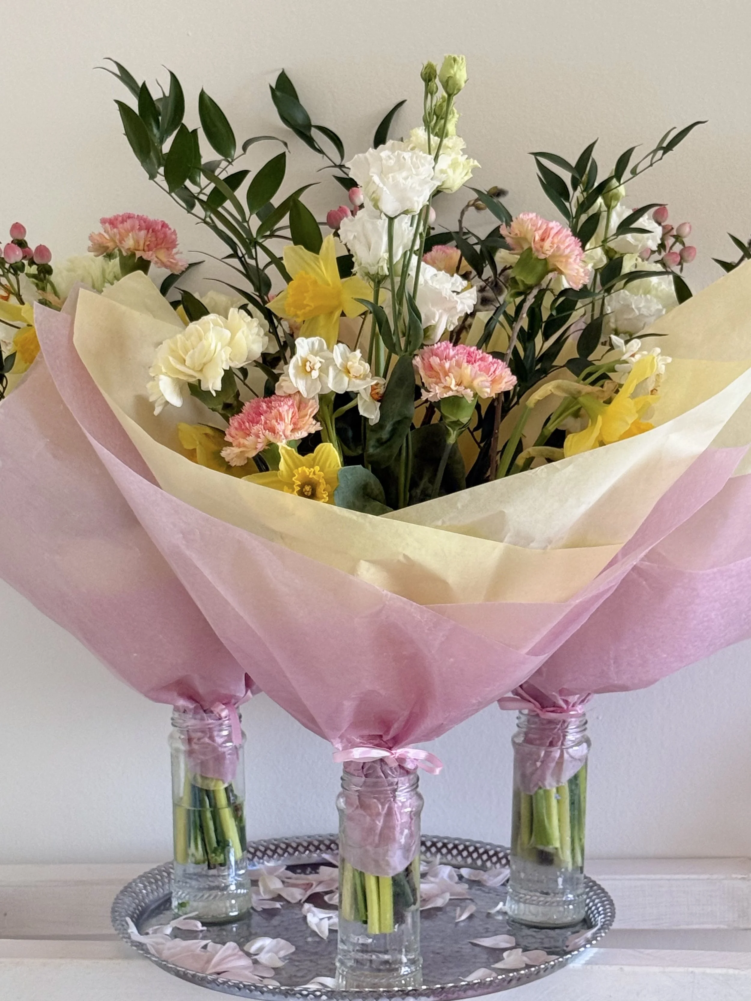 A floral arrangement with various flowers in pastel colors, wrapped in pink and cream tissue paper, displayed on three glass jars filled with water, placed on a decorative tray with petals scattered around.