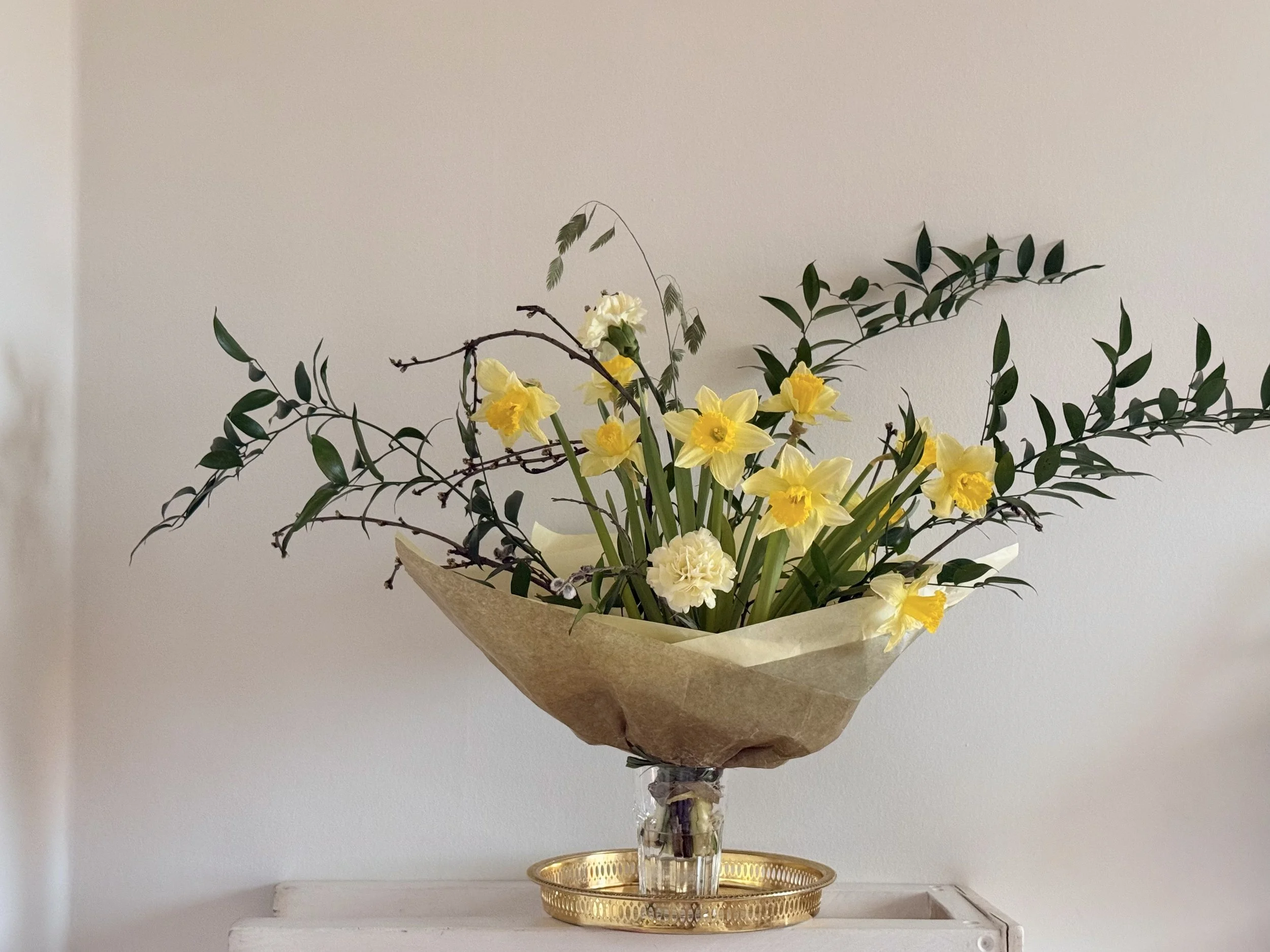 A bouquet of yellow and white flowers with green leaves and branches in a glass vase, wrapped in brown paper, placed on a gold tray on a white surface against a plain white wall.