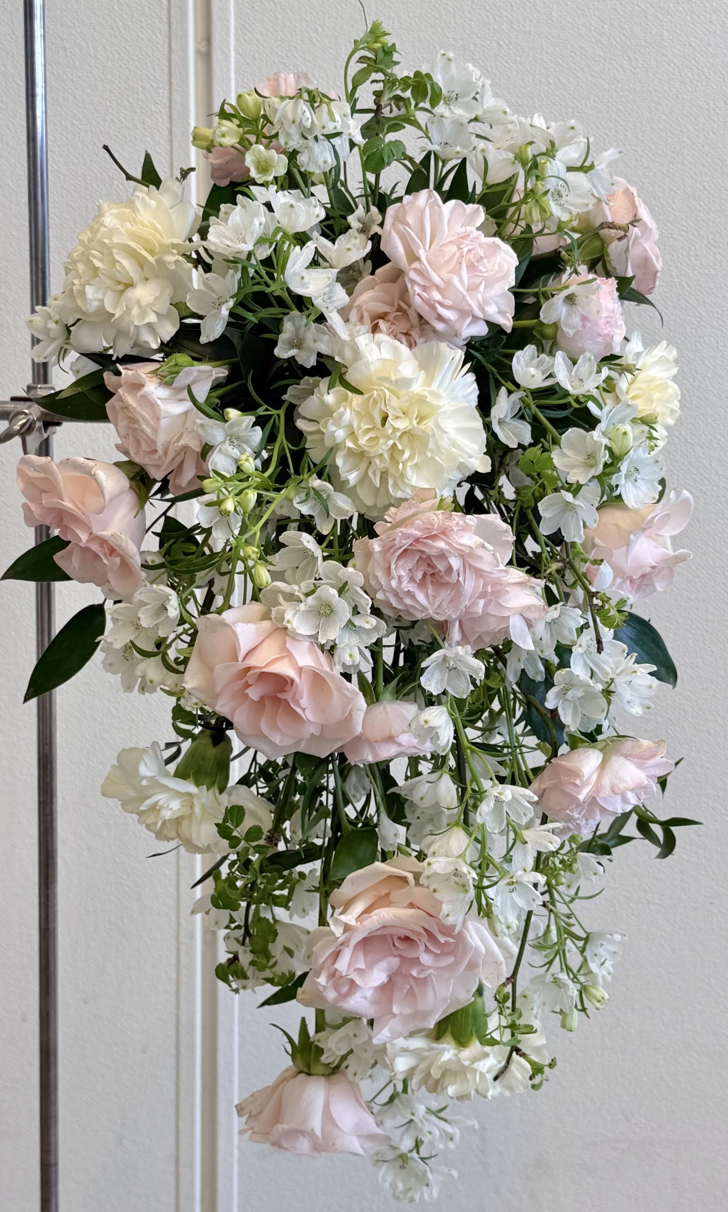A cascading arrangement of pale pink and white flowers, including roses, carnations, and other blooms, with green leaves against a plain background.
