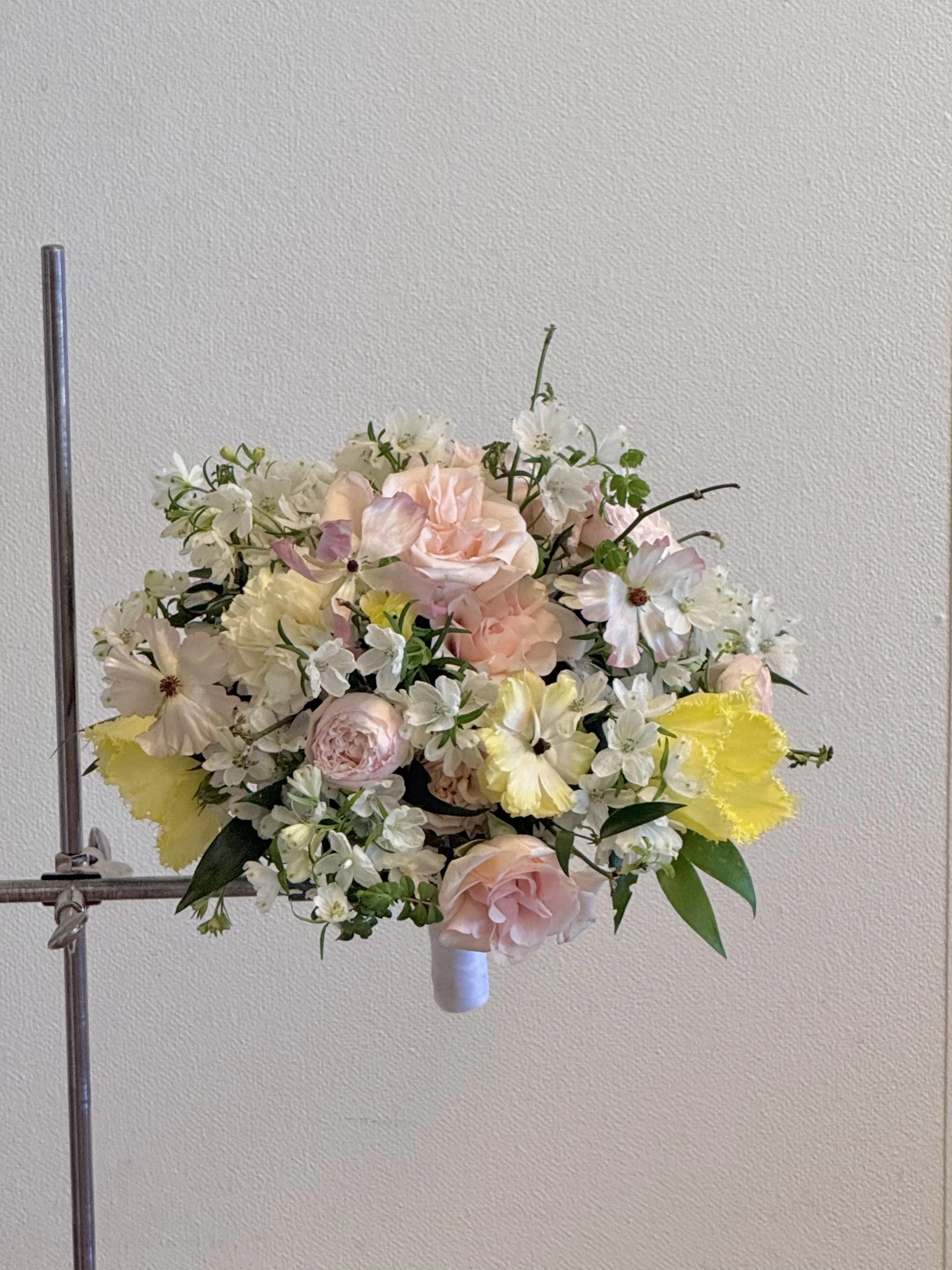 A floral arrangement with pink roses, white and yellow flowers, and green leaves, mounted on a white stand against a plain beige wall.