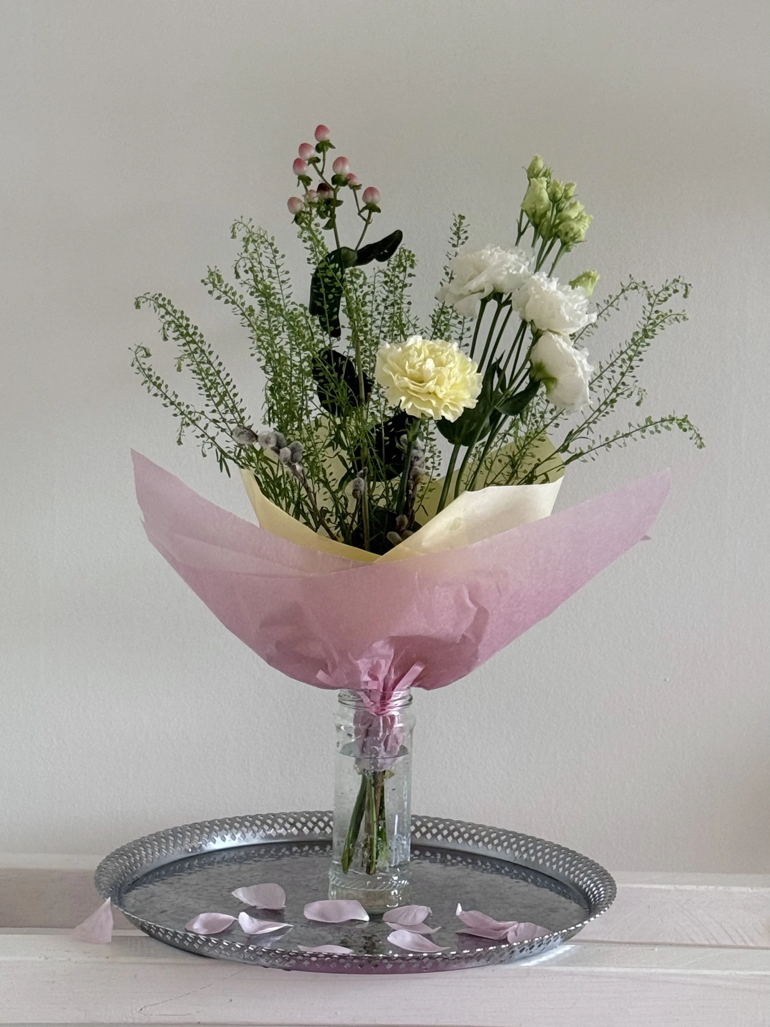 A flower arrangement with white and light pink flowers in a glass vase, wrapped in pink tissue paper, placed on a silver tray with pink and white flower petals scattered on it, against a plain white background.