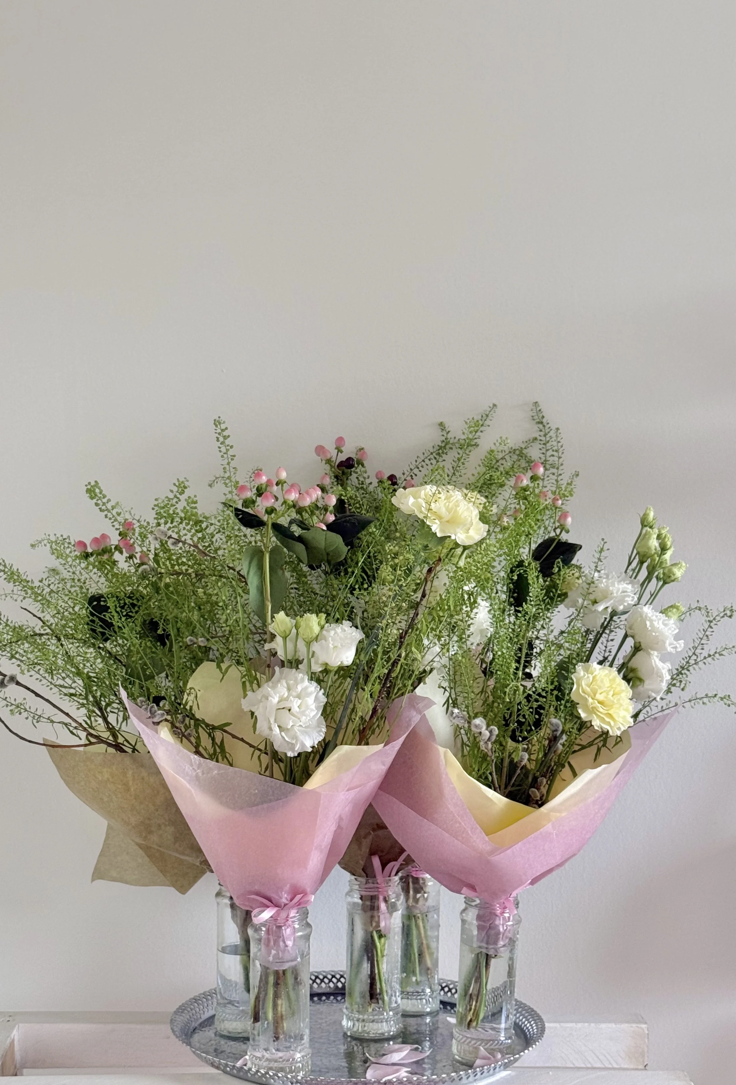 Three vases with colorful flowers and greenery, wrapped in pastel-colored paper, on a silver tray.
