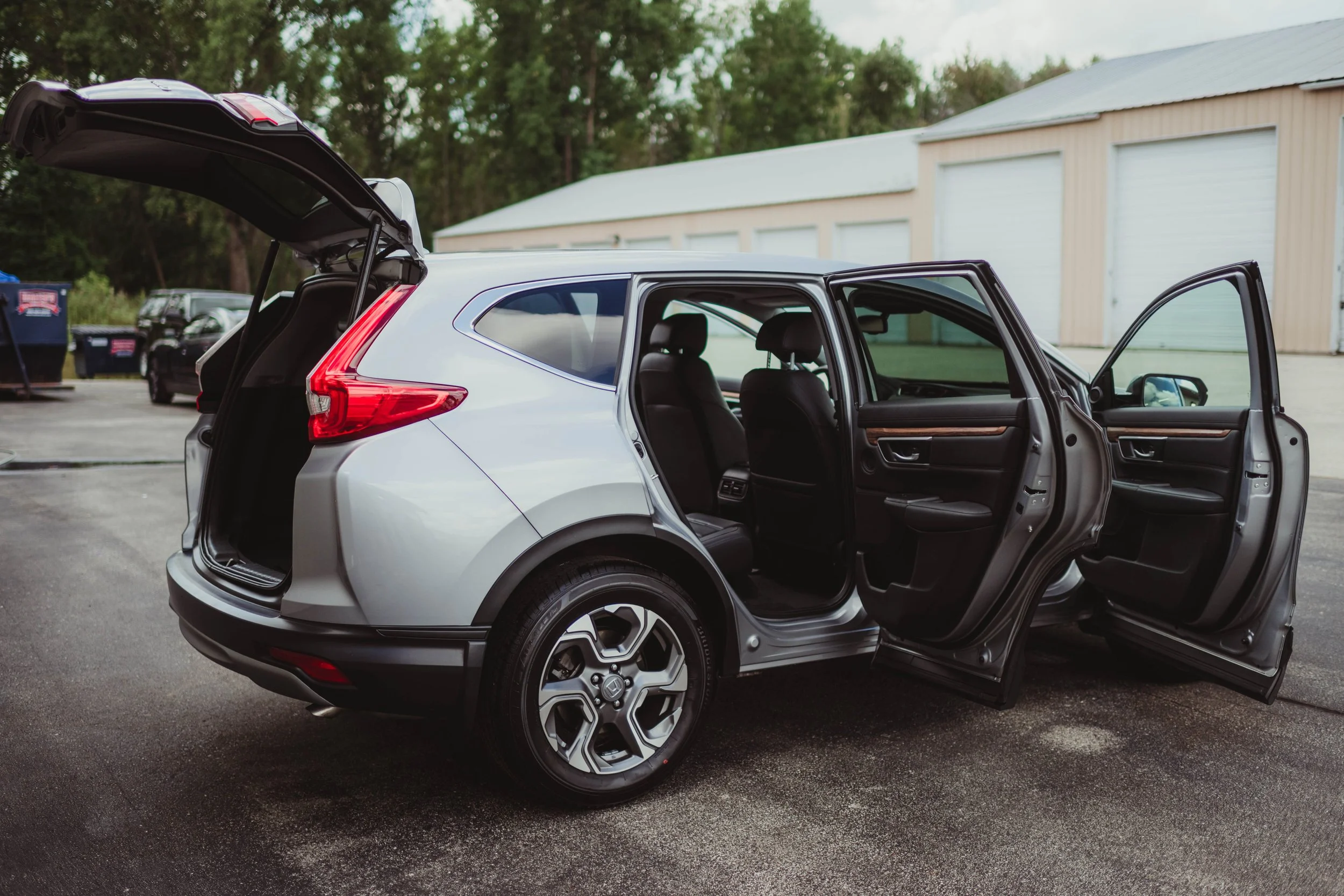 A silver SUV with its rear hatch open and front and rear doors open, parked outdoors near a building with a row of garages.