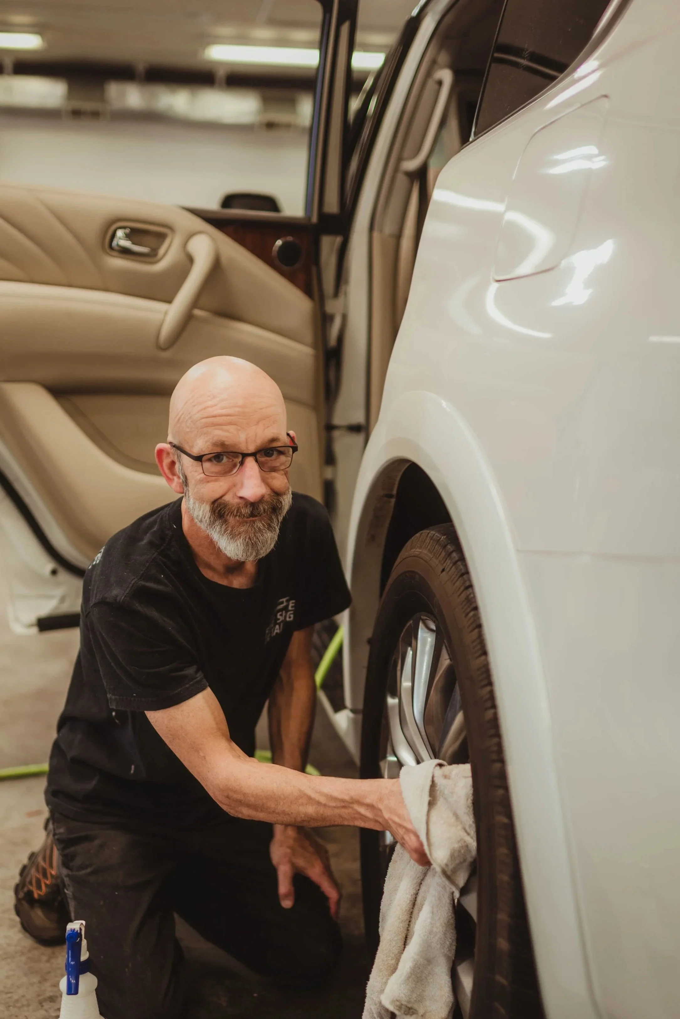 A man with glasses, a beard, and a black t-shirt is cleaning the tire of a white car using a white cloth in an indoor garage.