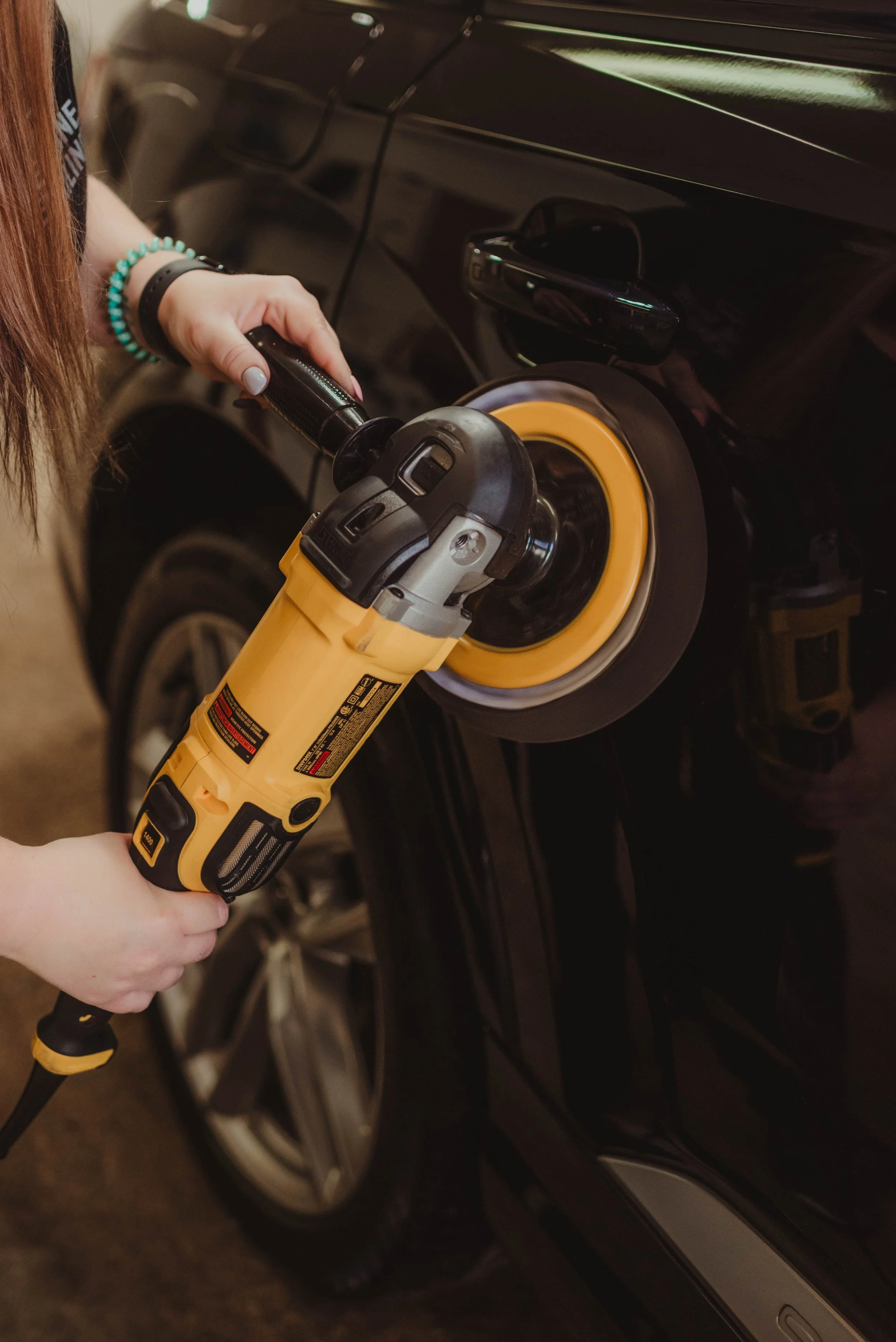 Person uses a yellow electric buffer to polish the black car's exterior.