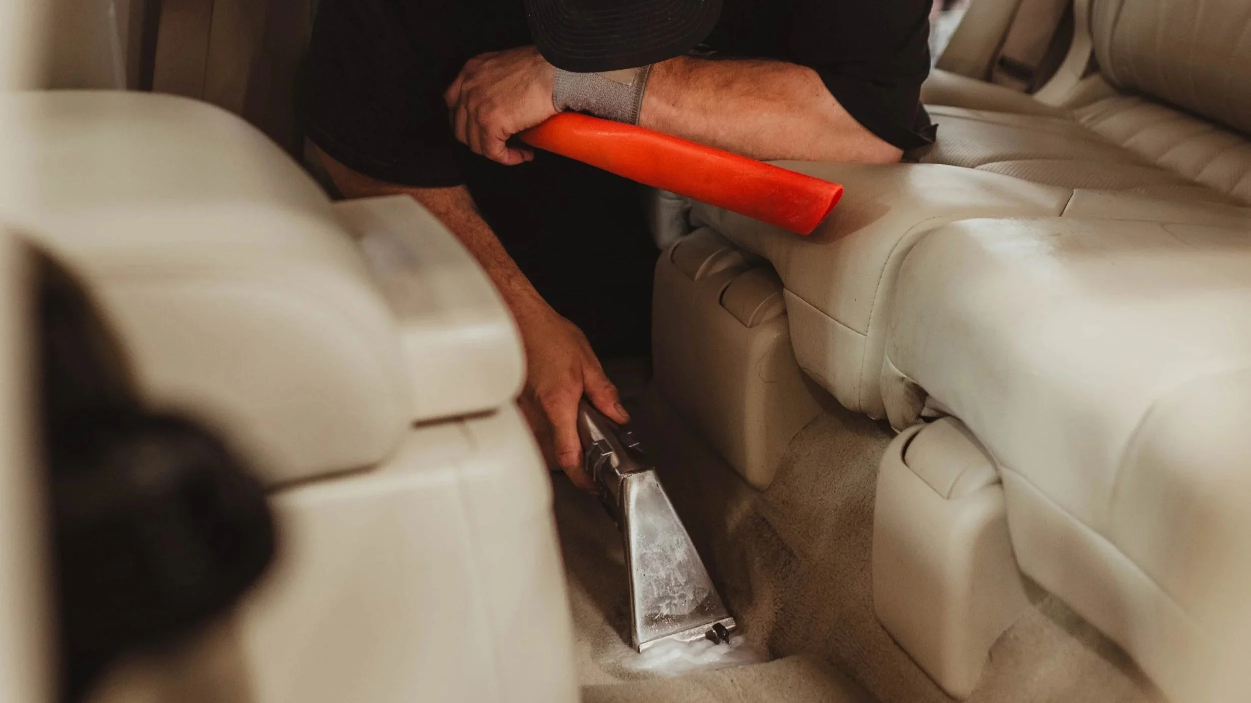 A person cleaning the carpeted floor of a beige leather car interior using a handheld vacuum cleaner while sitting on the seat with an orange pry bar resting on their arm.