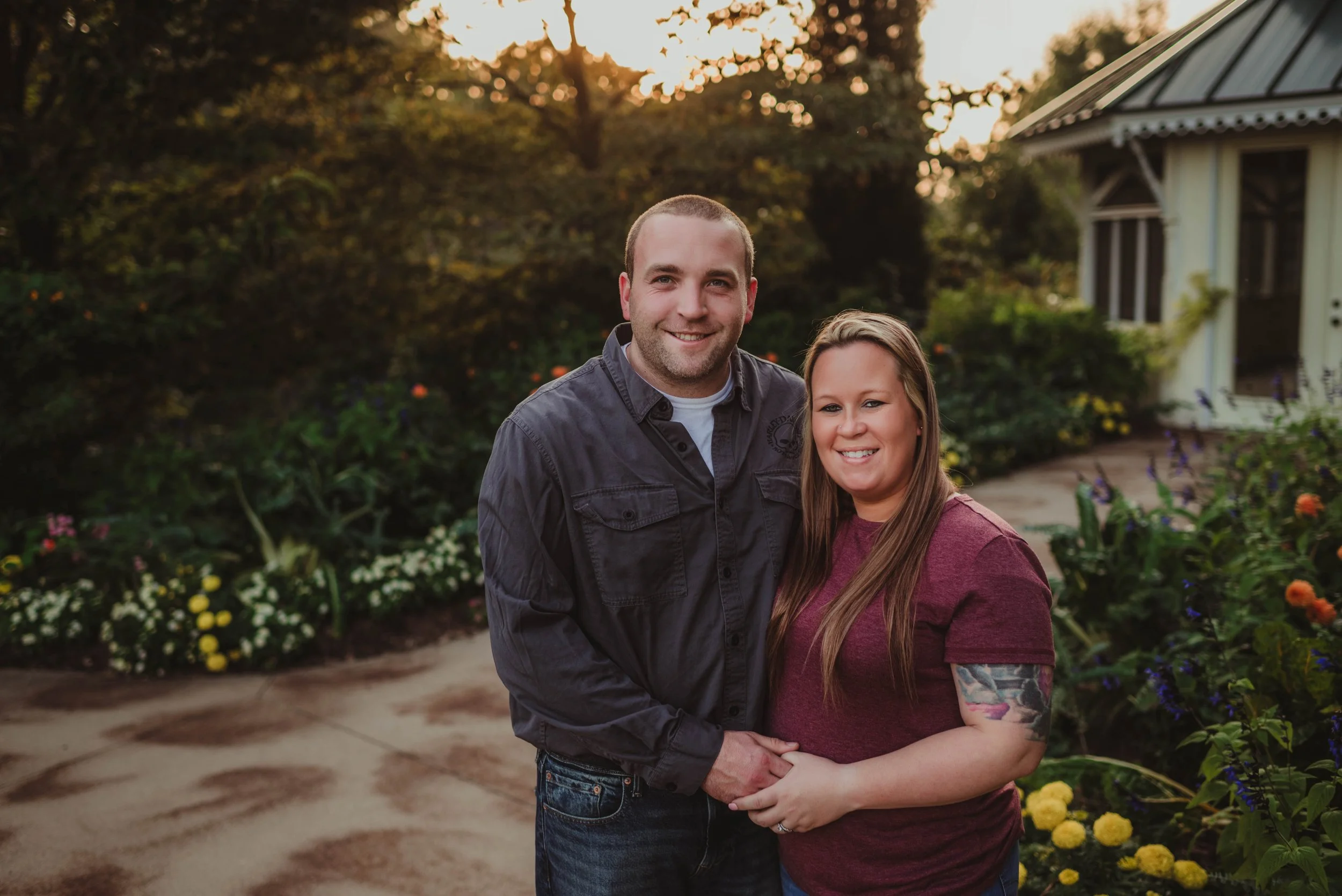A smiling couple stands hand in hand in a garden during sunset. The man wears a gray button-up shirt, and the woman has long hair, a tattoo on her right arm, and is wearing a maroon shirt. They are surrounded by colorful flowers and greenery, with a house visible in the background.