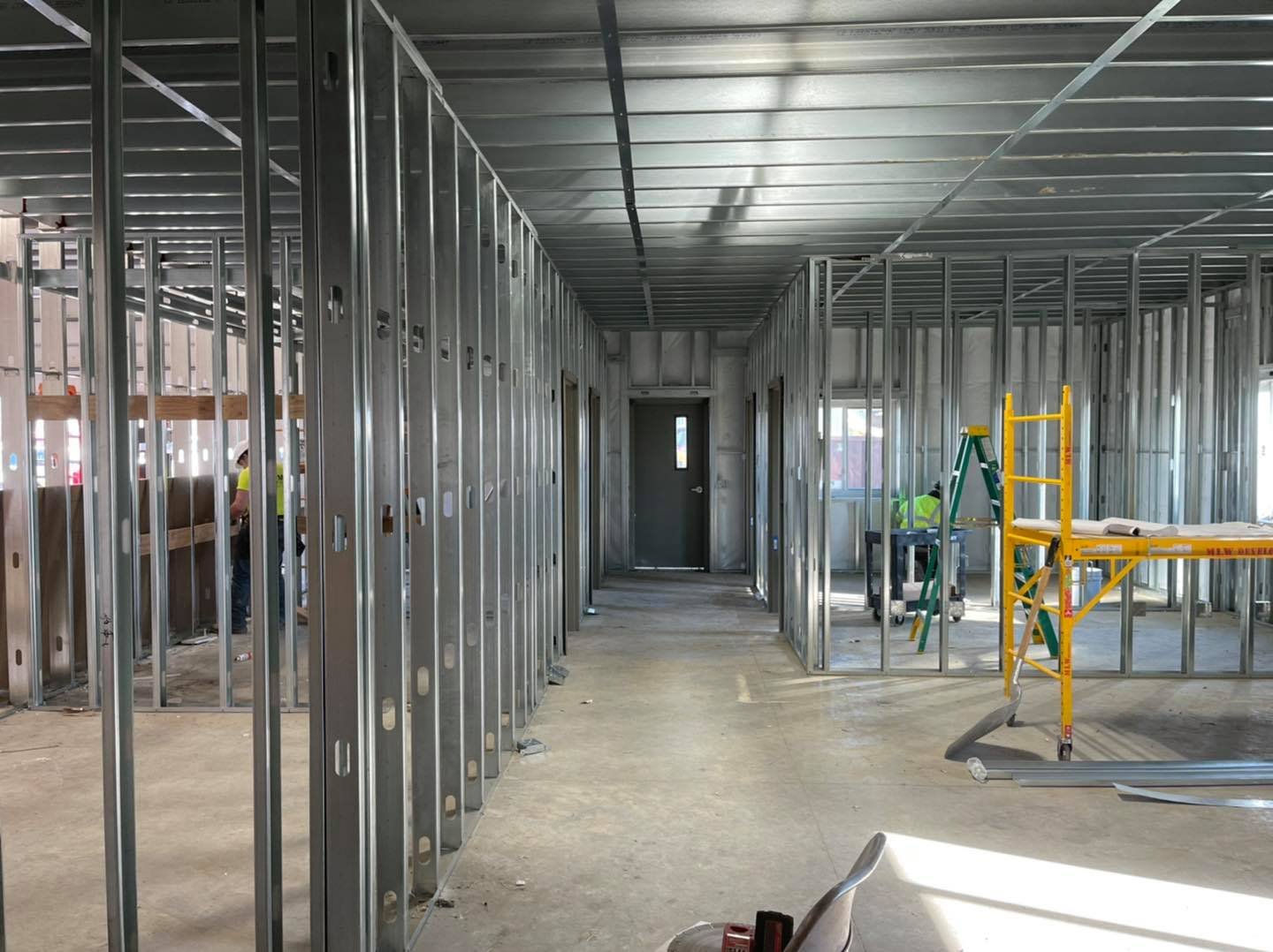 Interior of a building under construction with exposed metal framing, a ladder, and construction workers in safety vests.