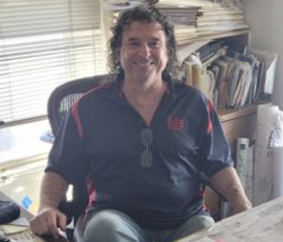A man with curly hair and glasses hanging from shirt collar, smiling, sitting at a desk in an office with books and papers in the background.
