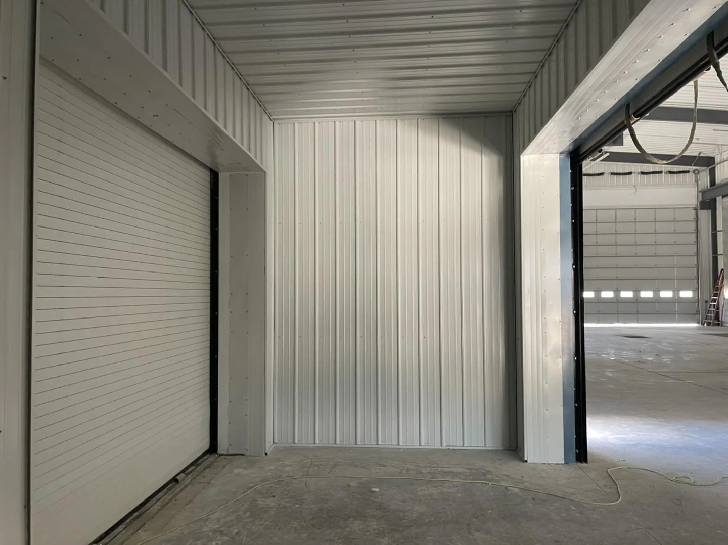 Interior view of a metal warehouse with two garage doors, one partially open, showing an industrial space with concrete floor and high ceiling.