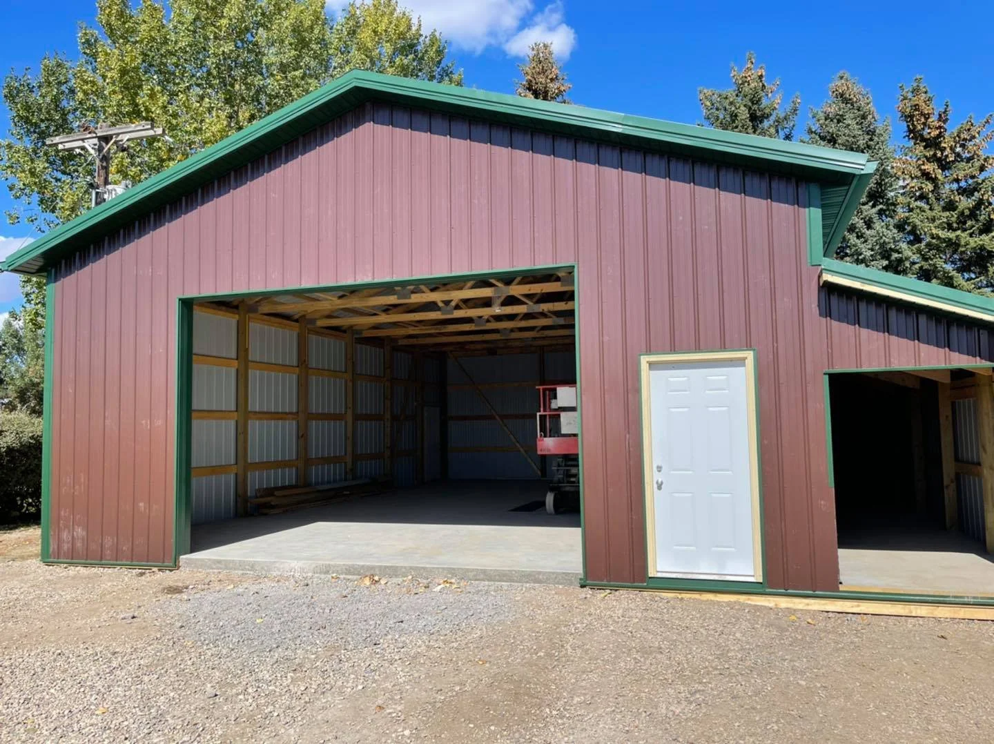 A barn under construction with a large open garage door, a side door, and green trim, surrounded by trees and a clear blue sky.