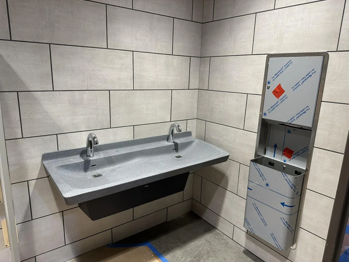 A public restroom with a double gray handwashing sink and a touchless soap dispenser or paper towel unit with a protective film, mounted on tiled beige walls.