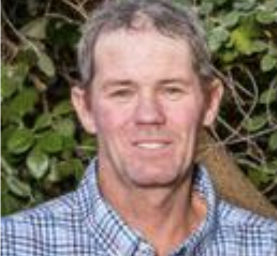 A middle-aged man with short gray hair wearing a blue checkered shirt stands outdoors in front of green foliage, smiling at the camera.