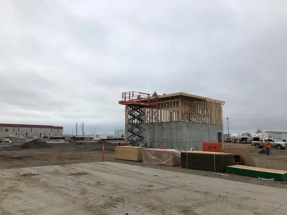 Construction site with a building in framing stage, heavy equipment, and workers in safety vests under a cloudy sky.