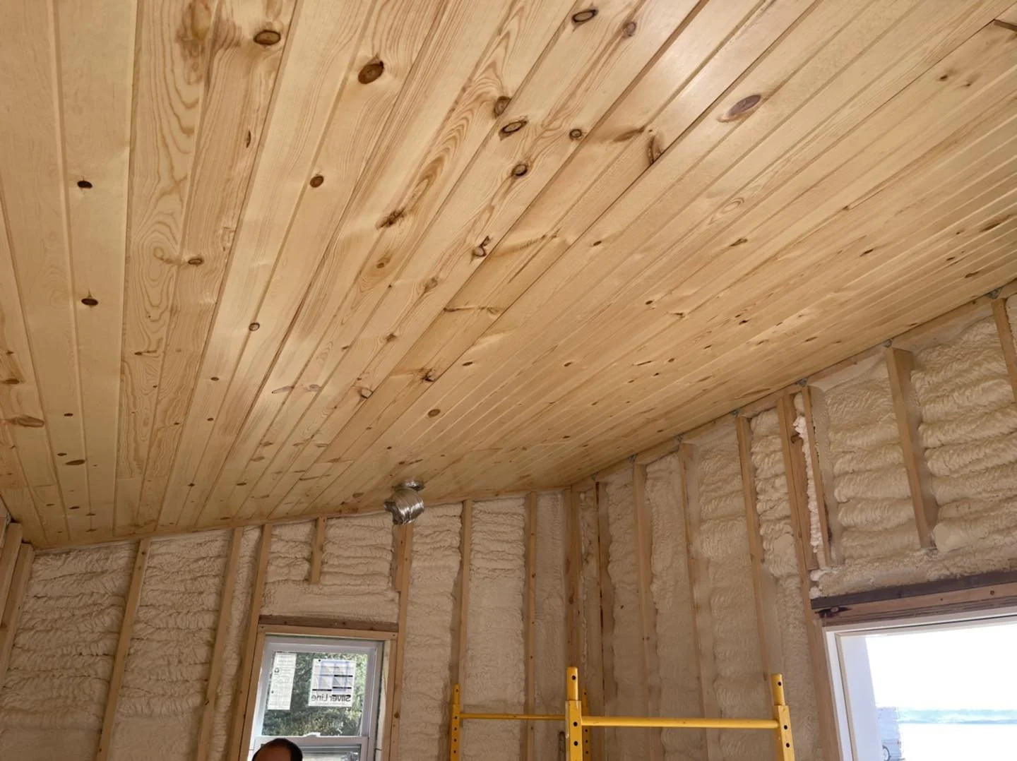 Interior of a room under construction with exposed insulation and wooden panel ceiling, scaffolding in the corner, and windows with view outside.
