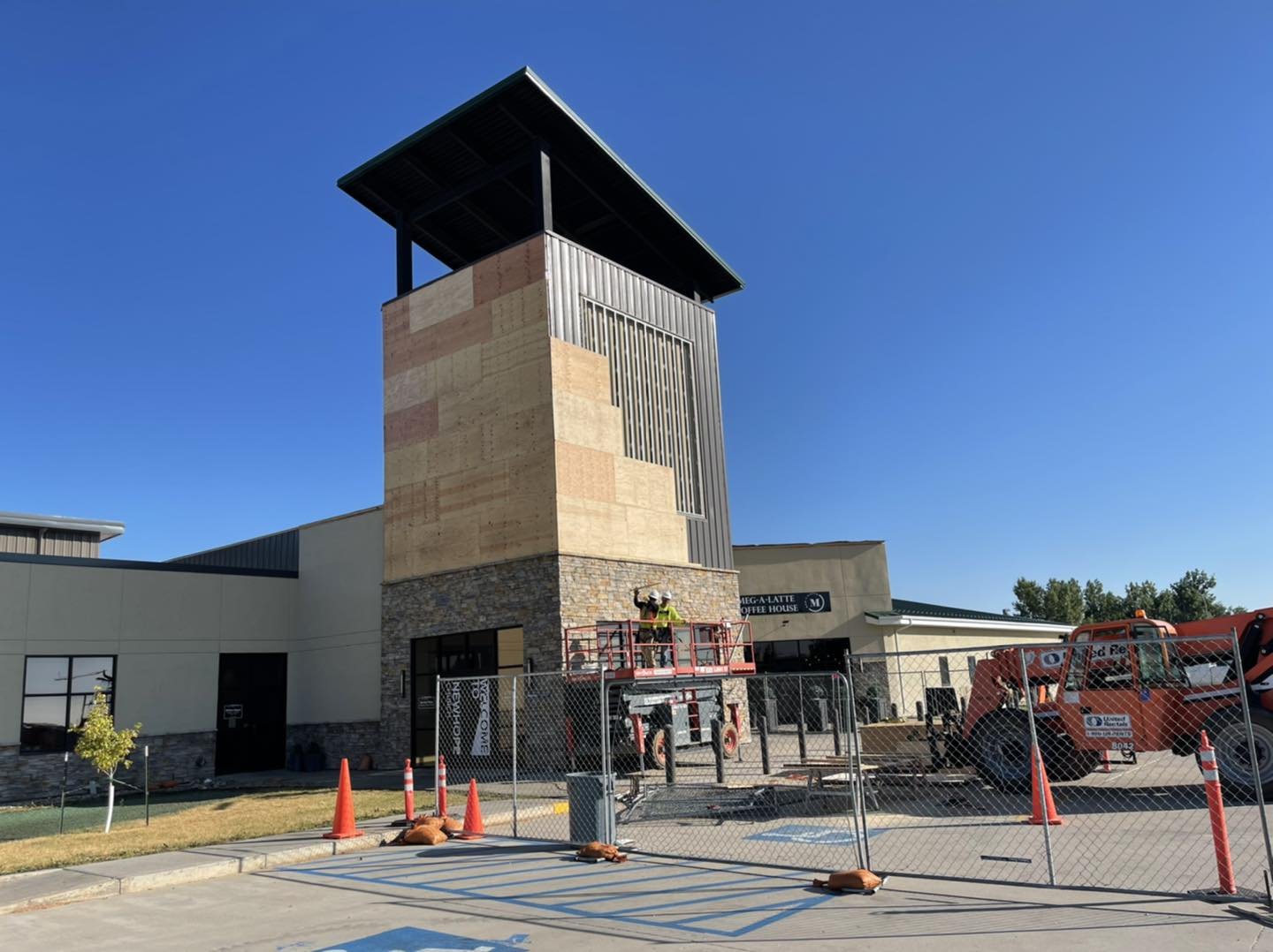Construction workers on a lift working on the exterior of a building with a tall tower, fenced-off construction site, and clear blue sky.